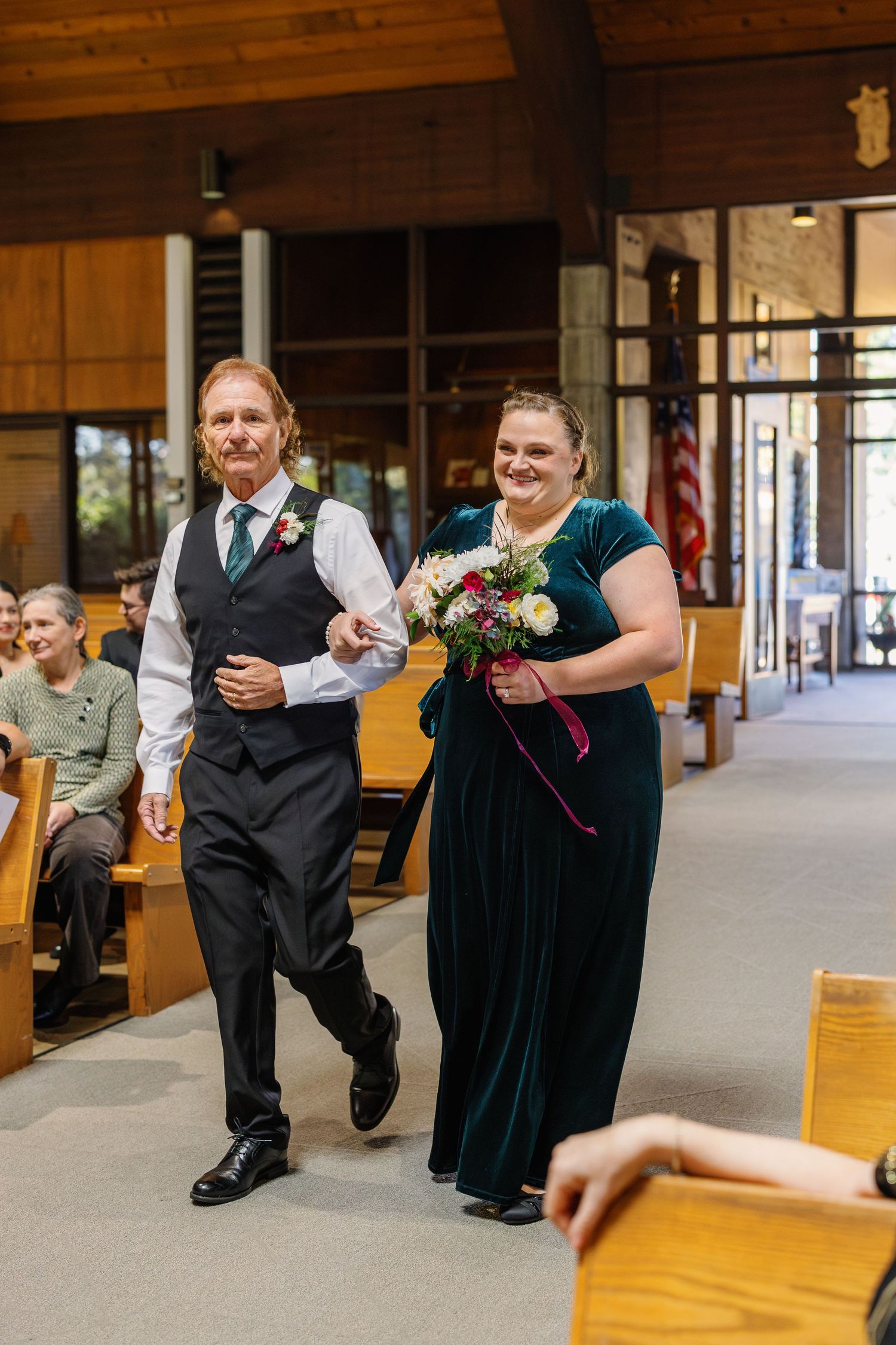Wedding party members walk down the church aisle, with attendant wearing an emerald velvet gown.