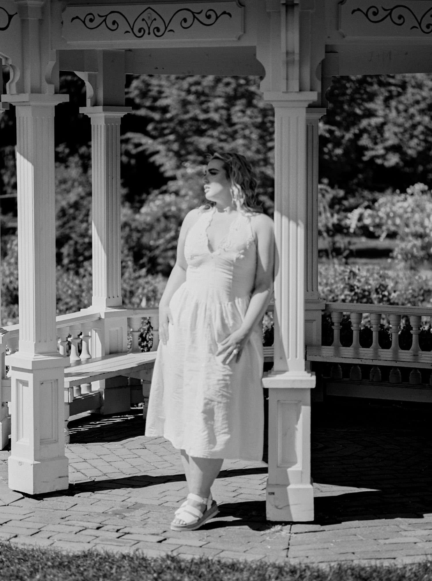 Woman in white dress standing in a sunlit gazebo, black and white photography.