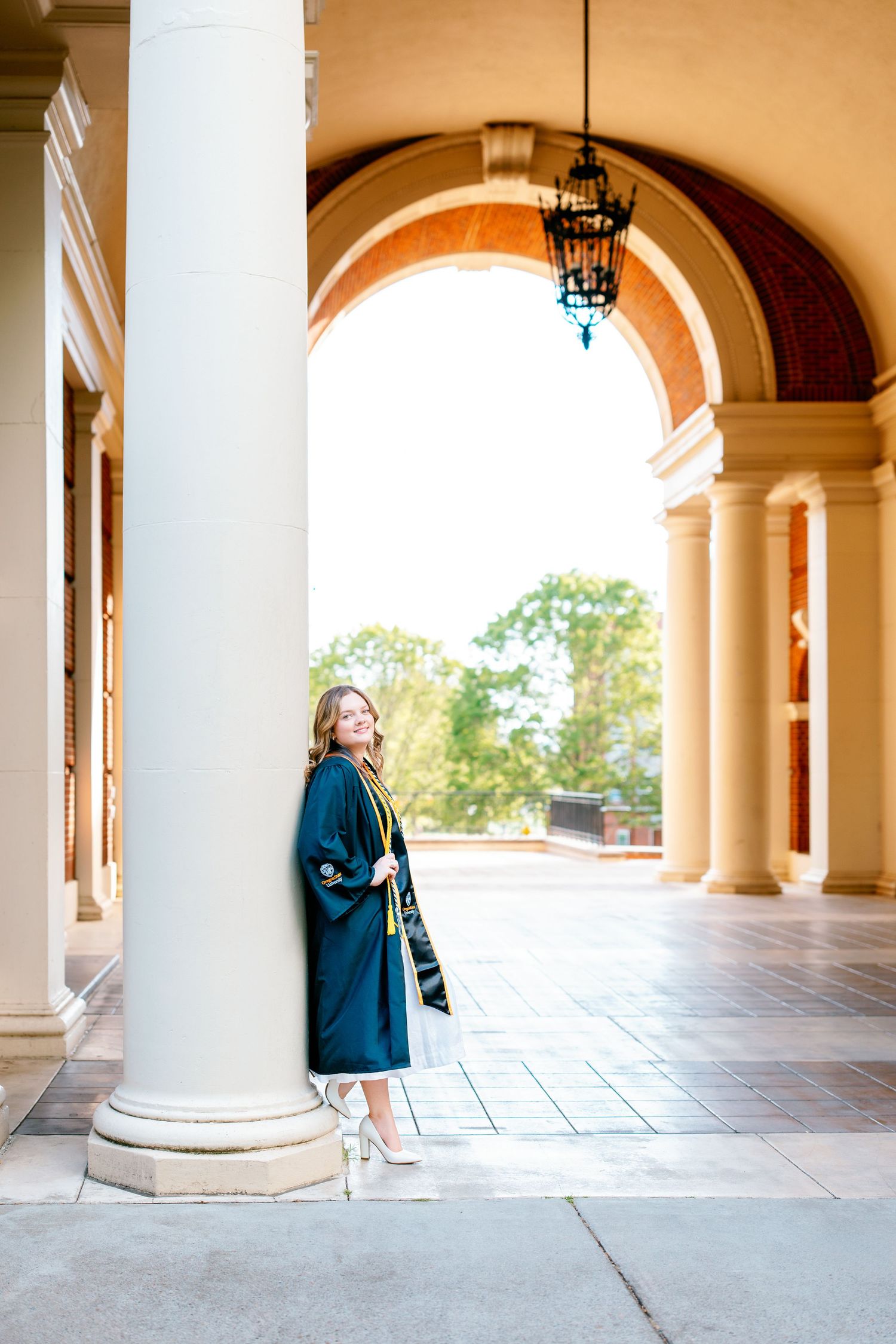 Graduate in teal cap and gown leaning against white column under arched colonnade at university campus.