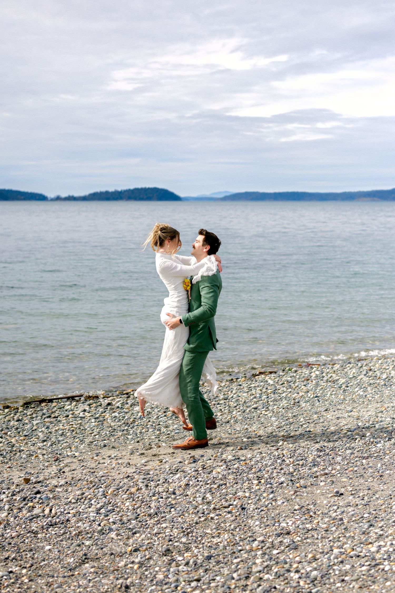 Groom in green suit lifts bride in white dress on rocky beach shoreline with calm ocean water behind them.