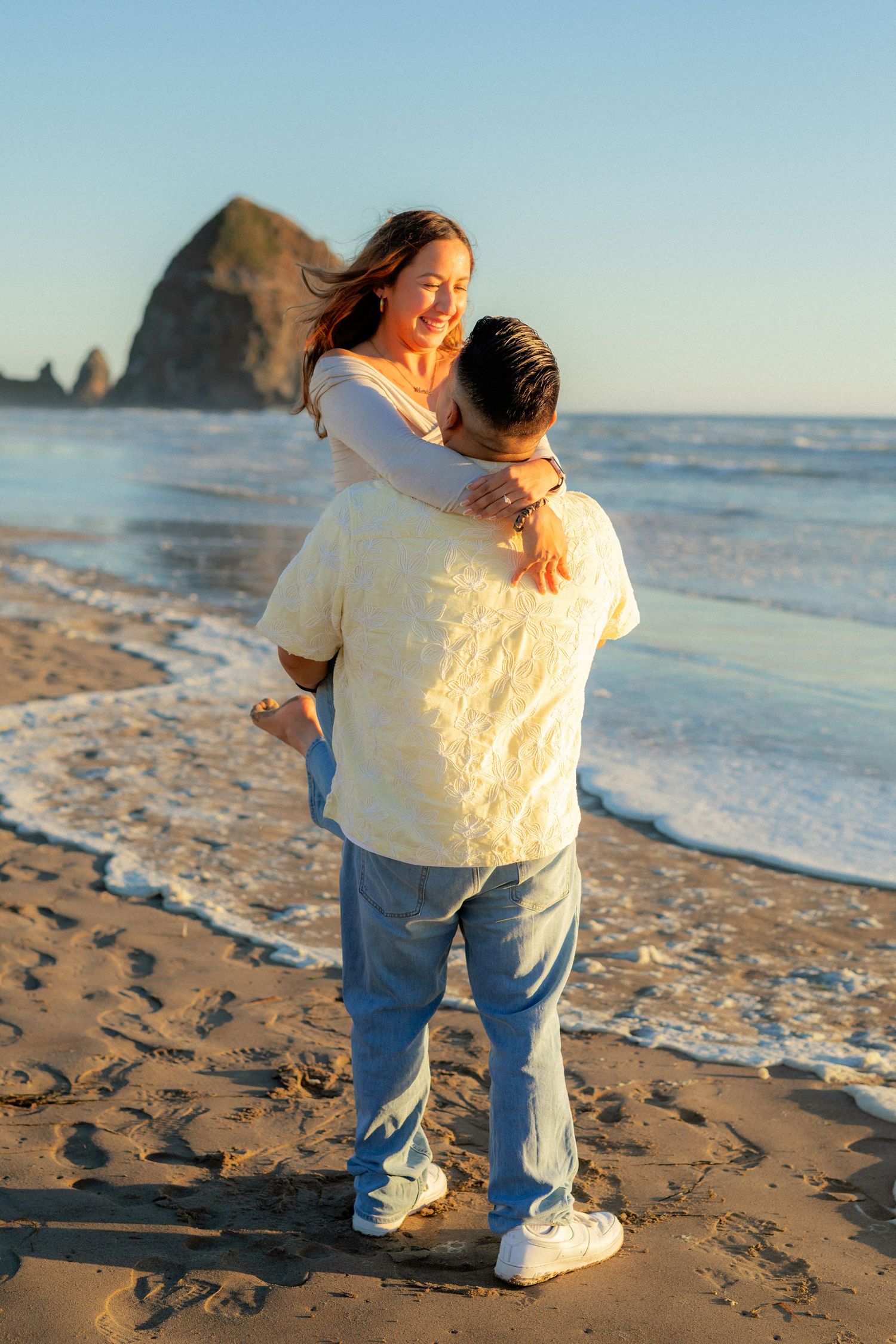 Couple embracing on sandy beach at golden hour with ocean waves and rocky sea stack in background.