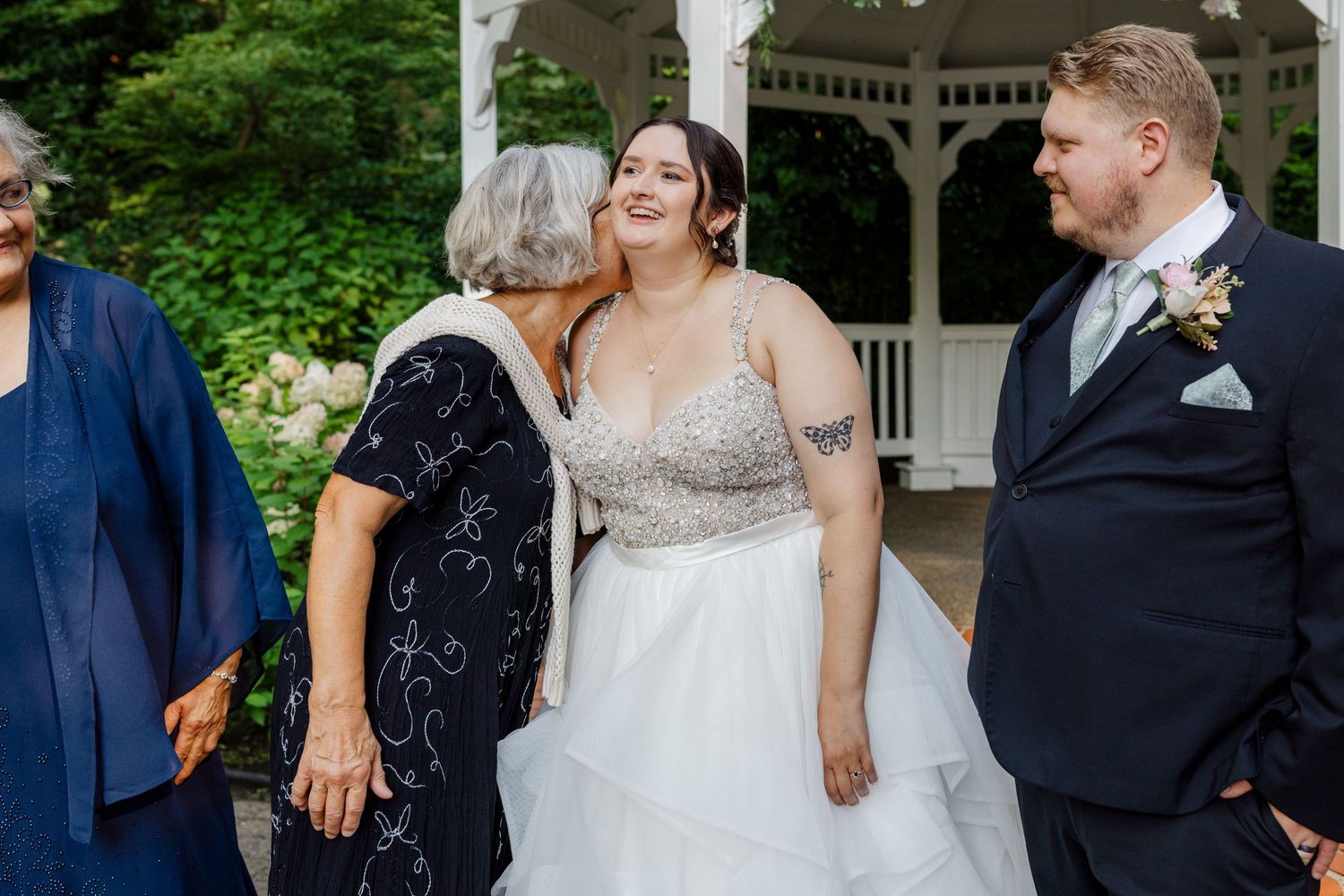 Bride in beaded gown receives kiss from elderly woman near gazebo as groom in suit stands nearby.