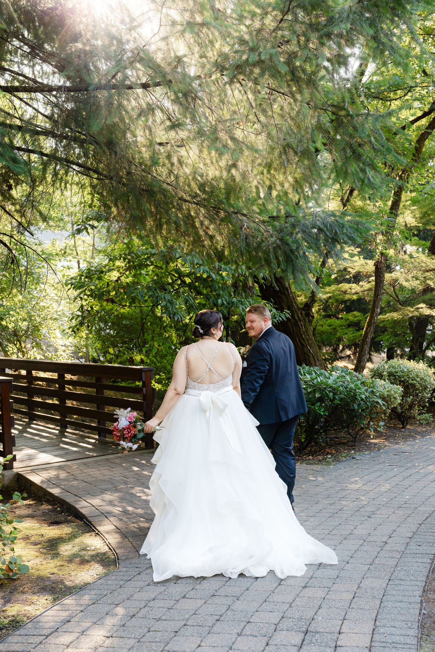 Bride in white gown and groom in navy suit walk hand-in-hand along a garden path at their wedding.