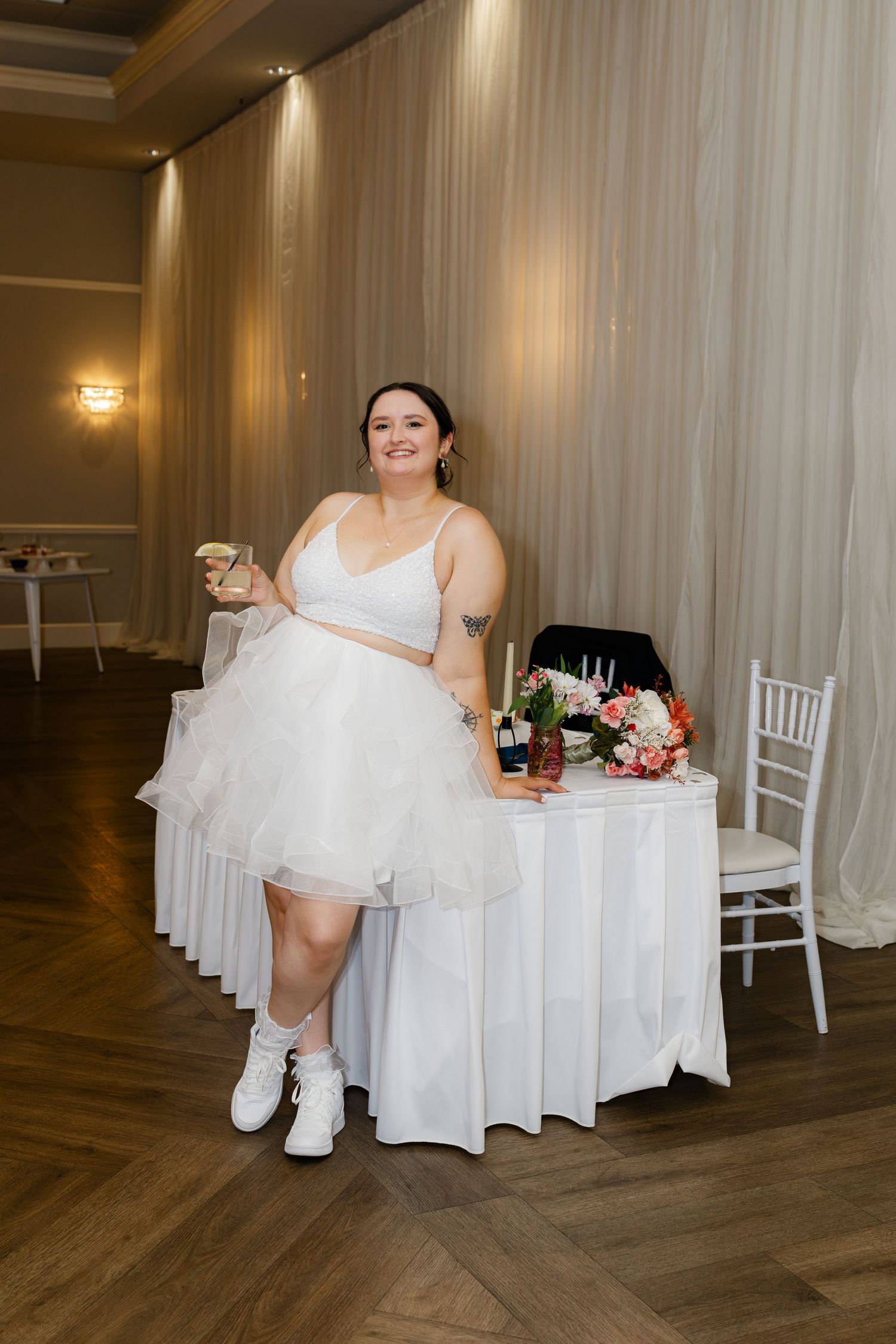 Bride in short white dress and sneakers smiling while leaning against sweetheart table at reception.