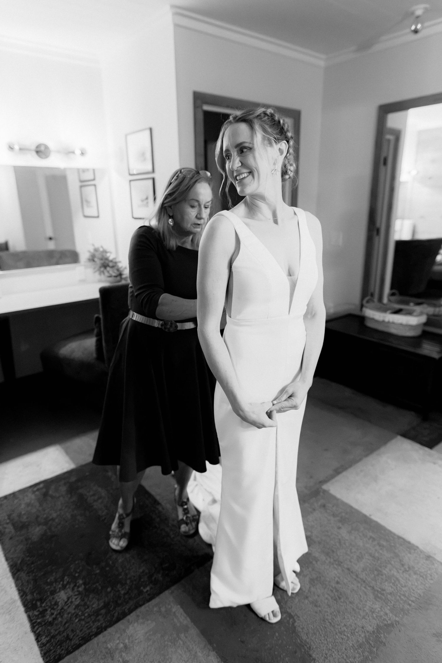 Black and white photo of a bride in a white gown smiling as a woman in black dress helps fasten her wedding dress.