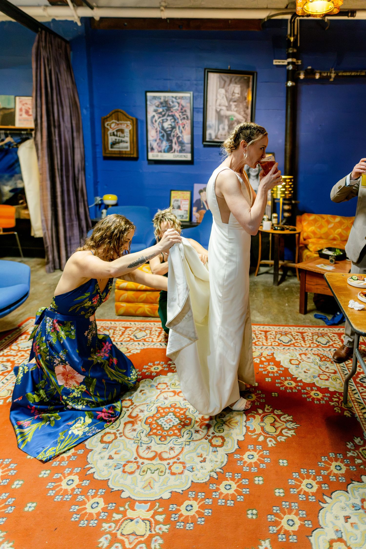 Bridesmaid in floral dress adjusts bride's white gown in colorful blue-walled room with orange patterned rug.