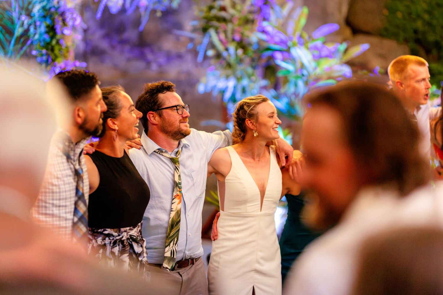Bride and groom embracing with guests during colorful lit wedding reception.