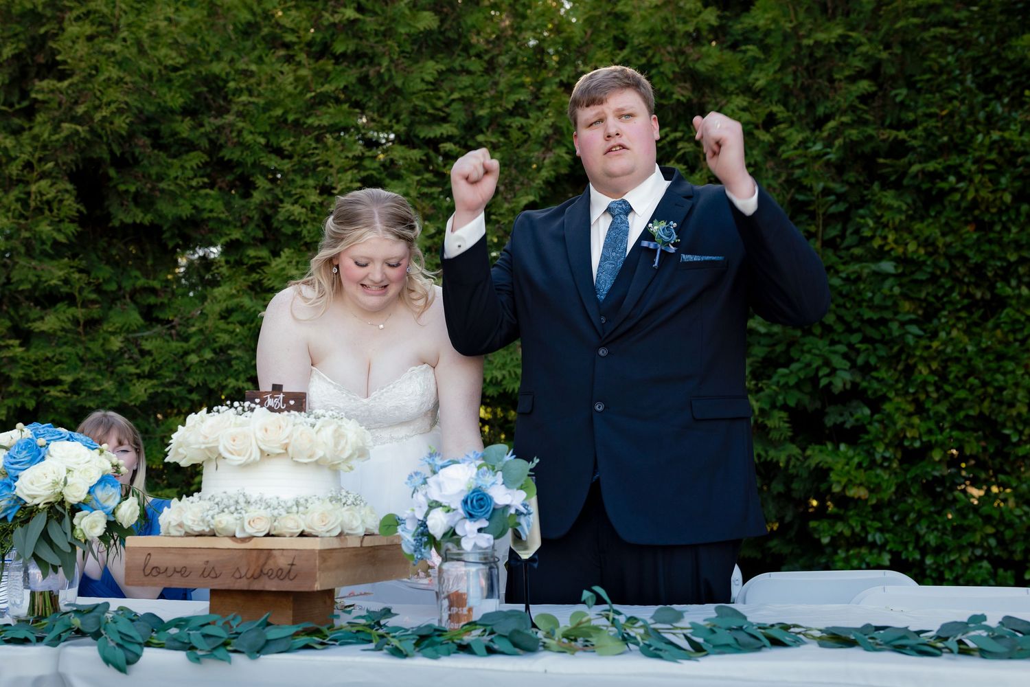 Groom celebrating with raised fists beside bride at outdoor wedding reception table with floral arrangements.