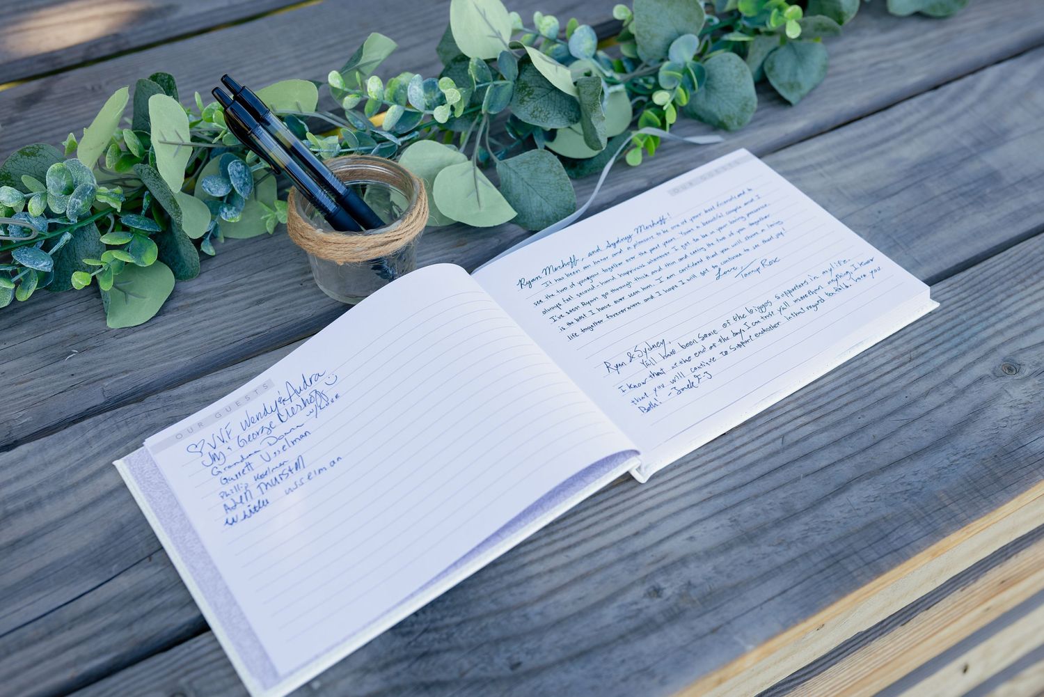 Open guest book with handwritten messages on wooden table, decorated with eucalyptus greenery and a pen holder.