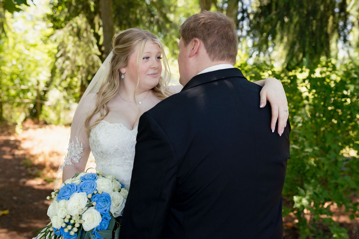 Bride in lace gown embraces groom outdoors, holding a blue and white floral bouquet in a wooded setting.