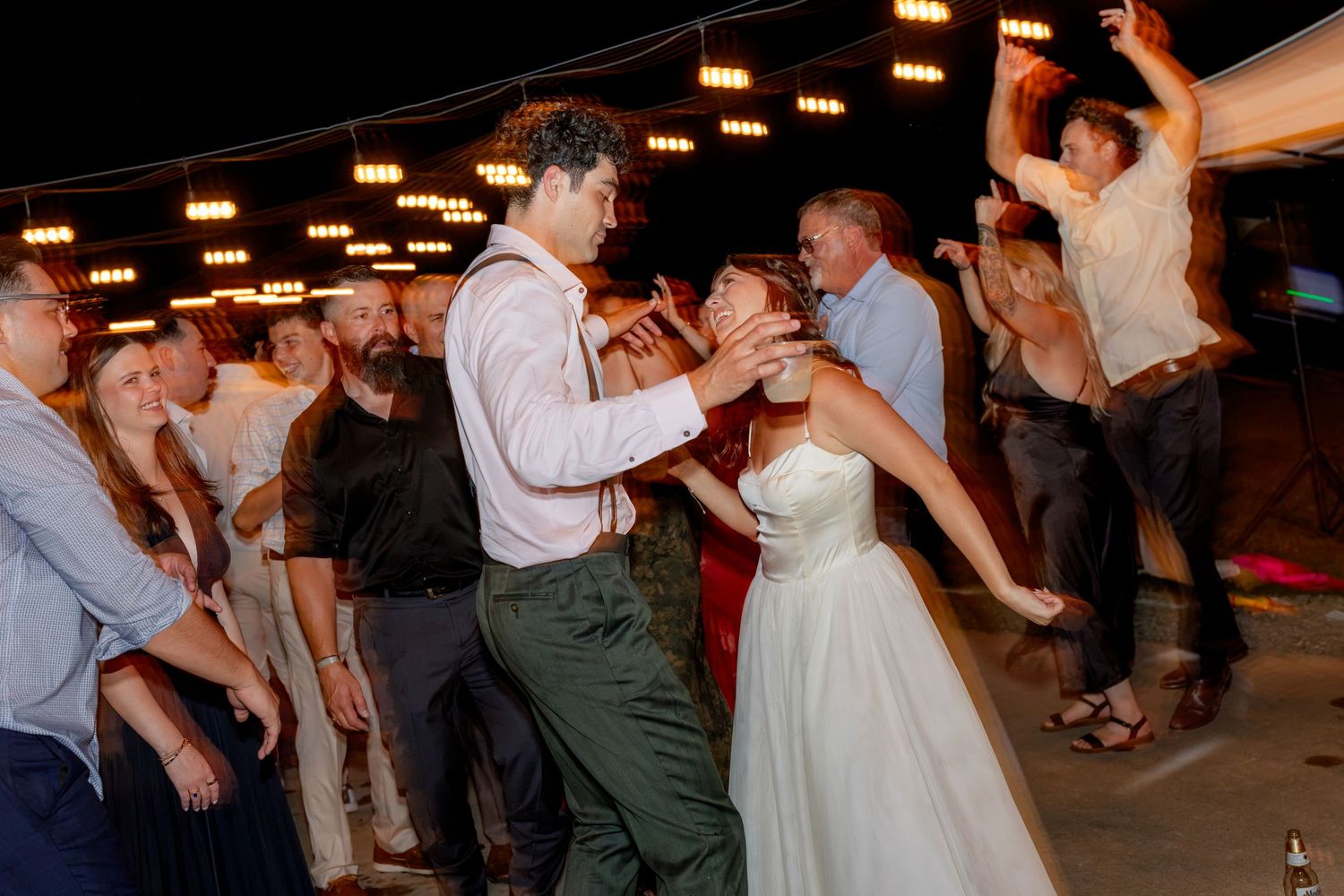 Bride and groom dancing joyfully with guests at a lively wedding reception under warm string lights.