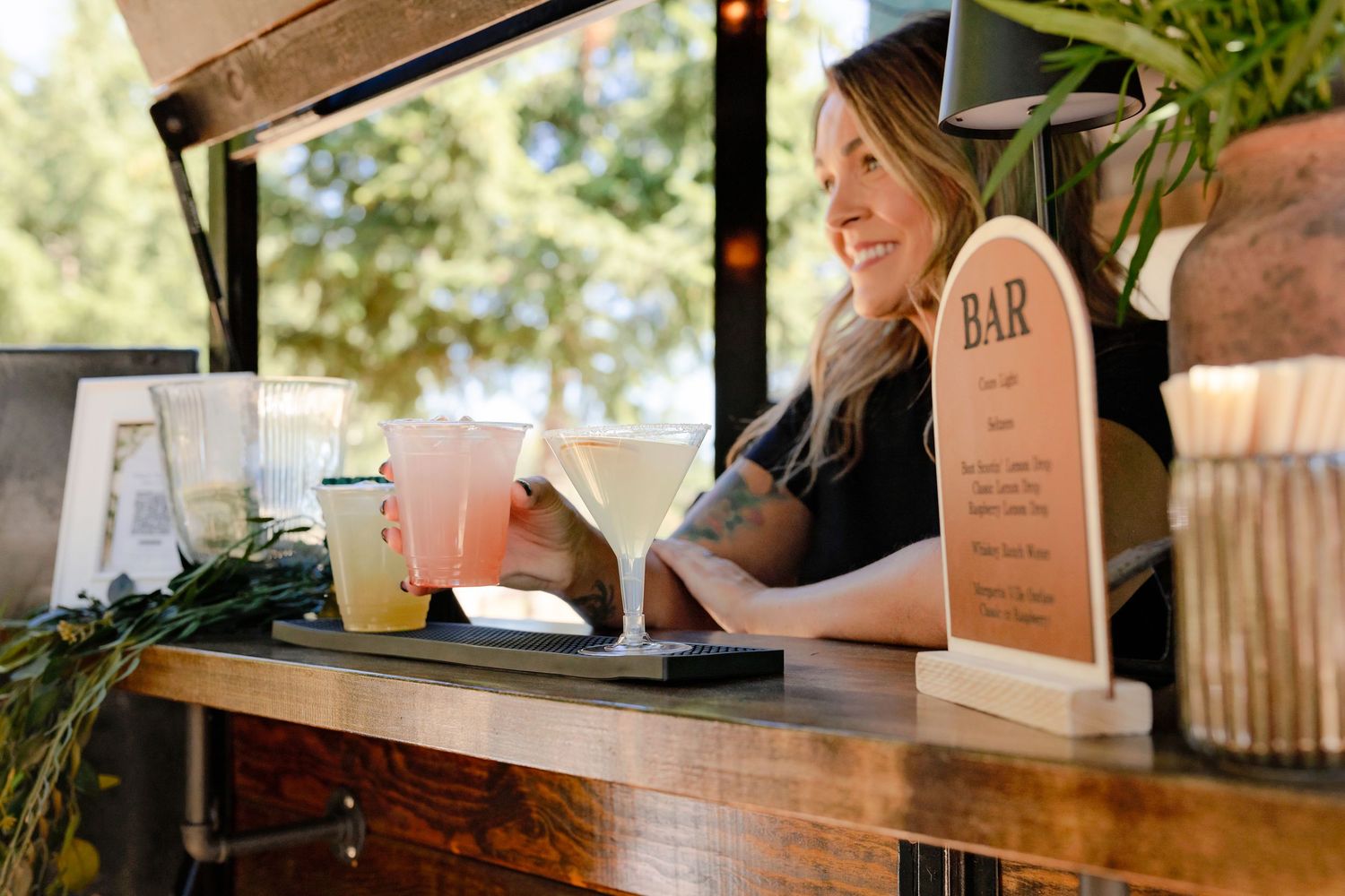 Smiling female bartender serving drinks at an outdoor bar counter with a rustic wooden finish.