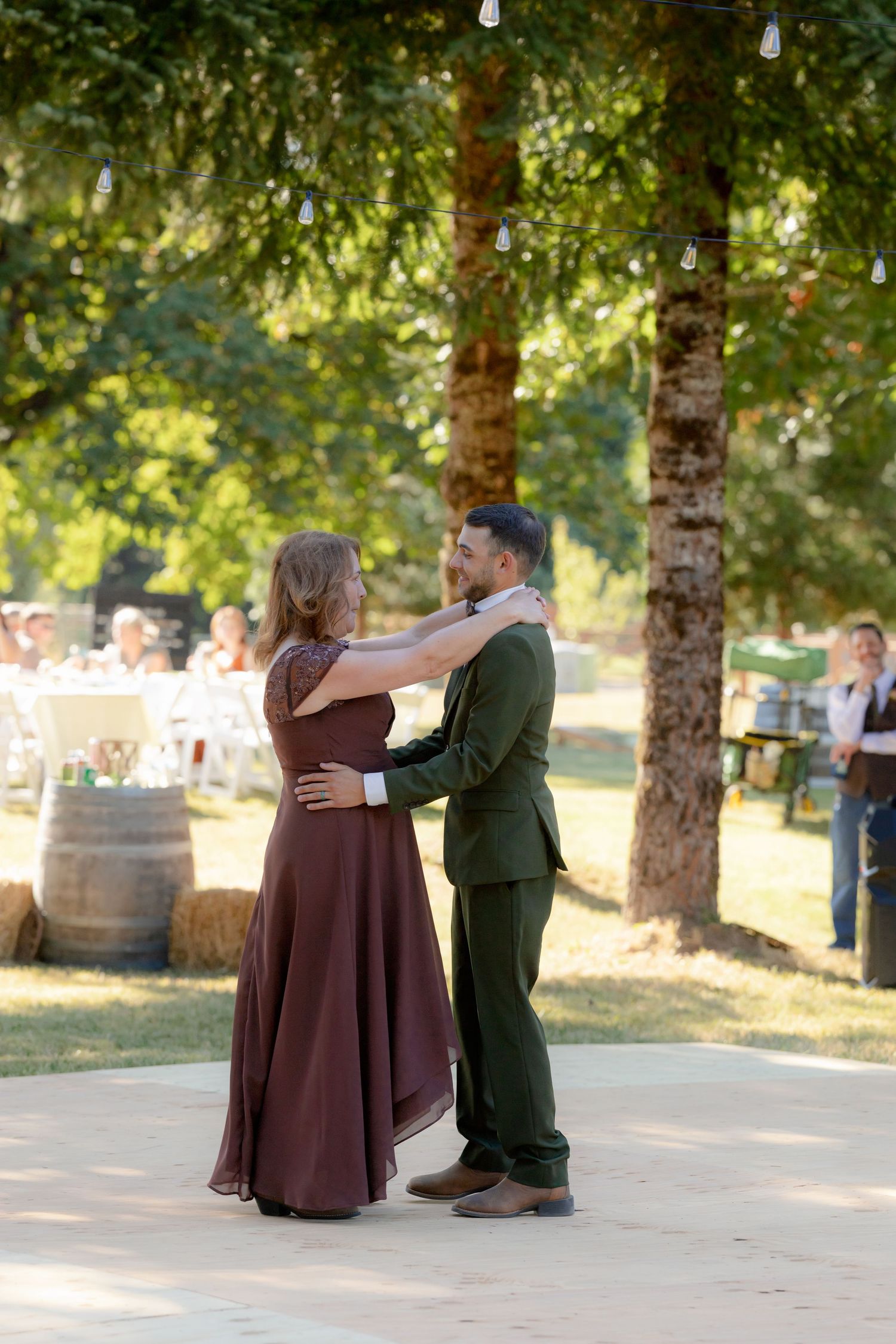 A couple shares a romantic outdoor dance at a rustic wedding reception surrounded by trees and string lights.