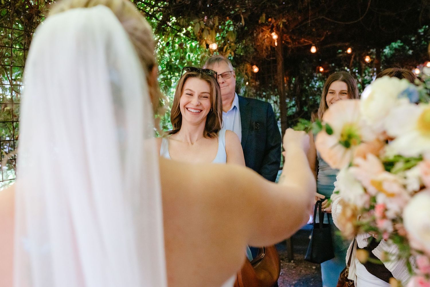A smiling bridesmaid faces the bride during an outdoor wedding ceremony surrounded by warm string lights.