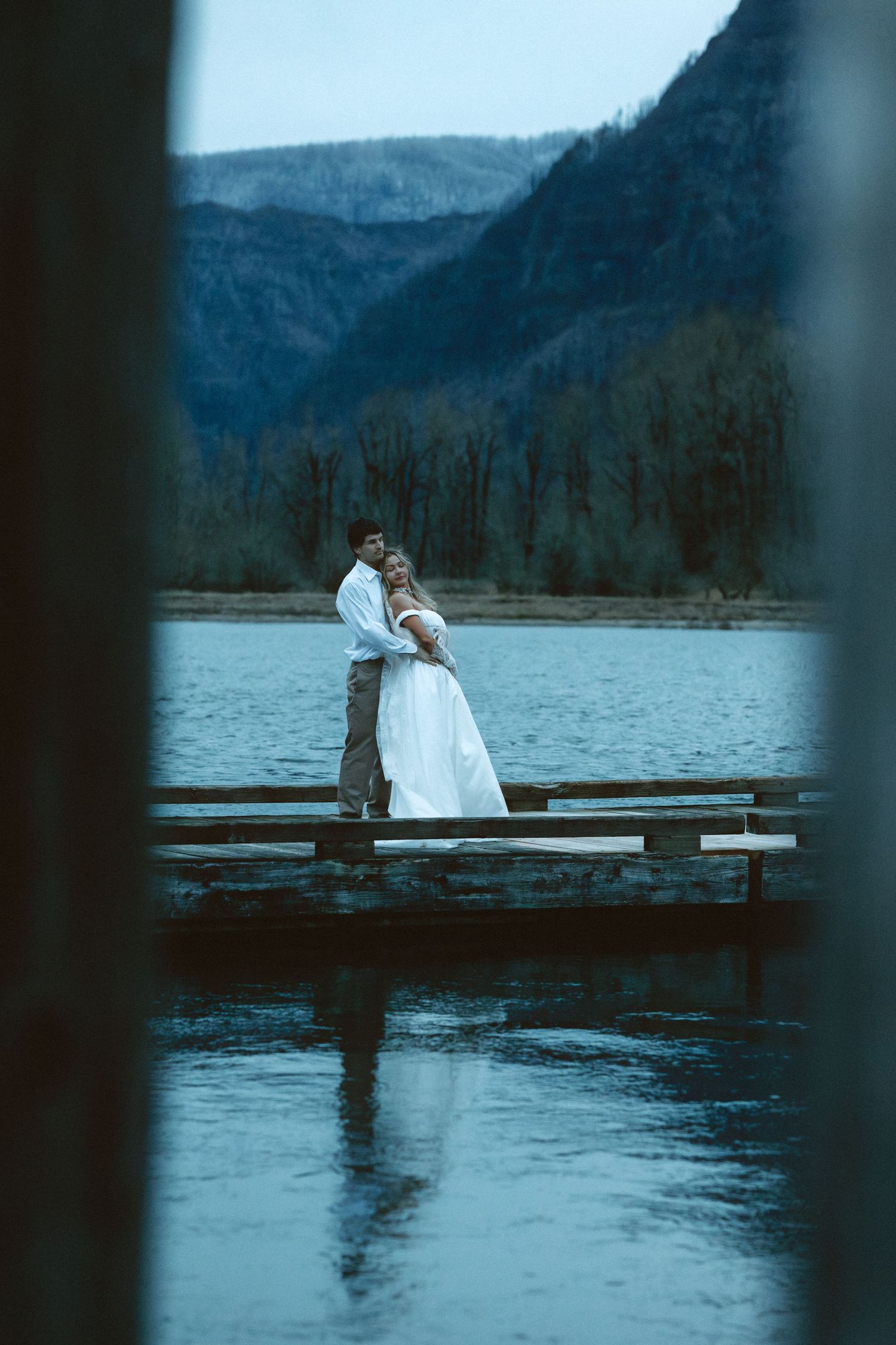 Couple standing together on a wooden dock overlooking a misty mountain lake at dusk.