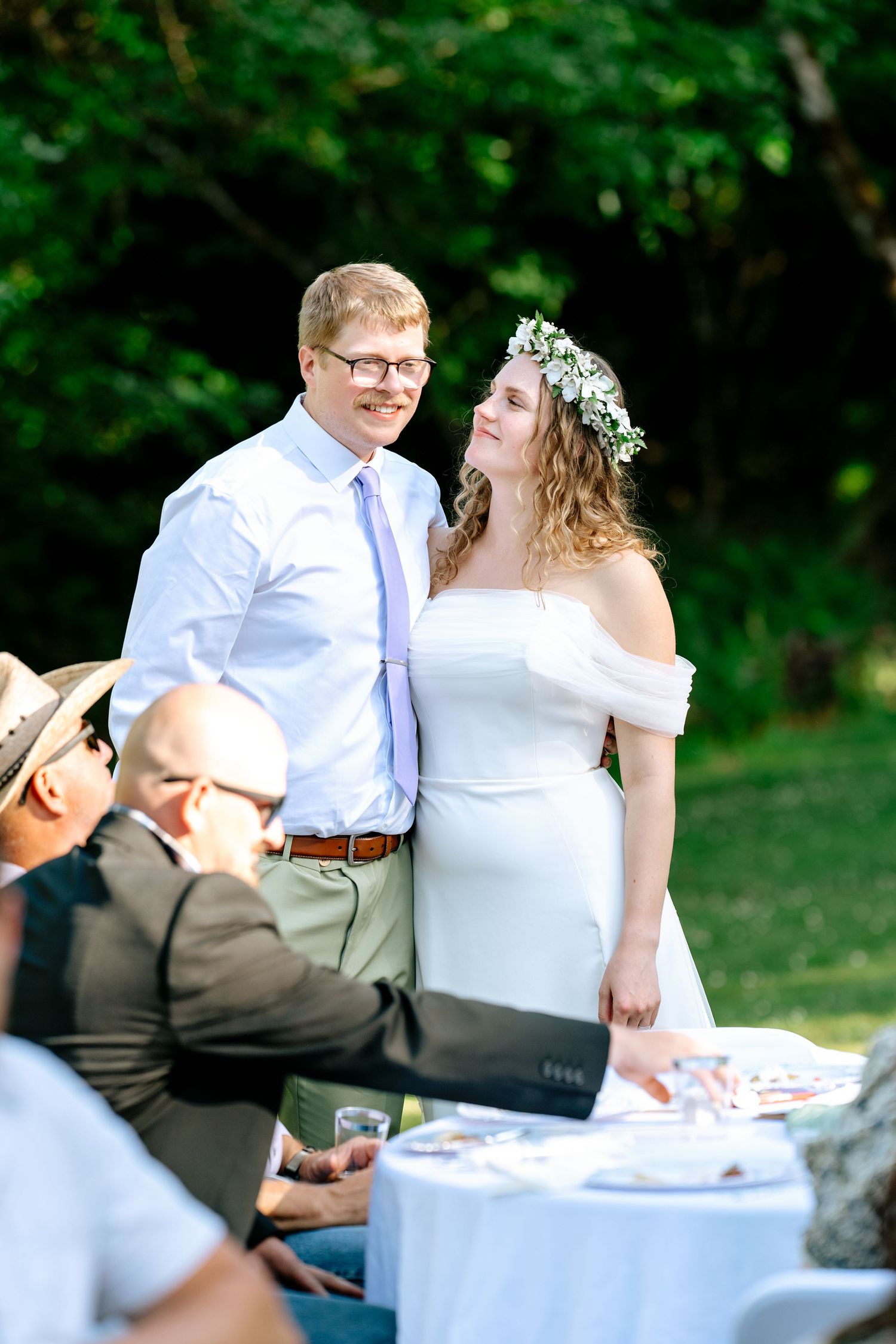 Smiling groom in white shirt and lavender tie stands beside bride in white dress with floral crown at outdoor reception.