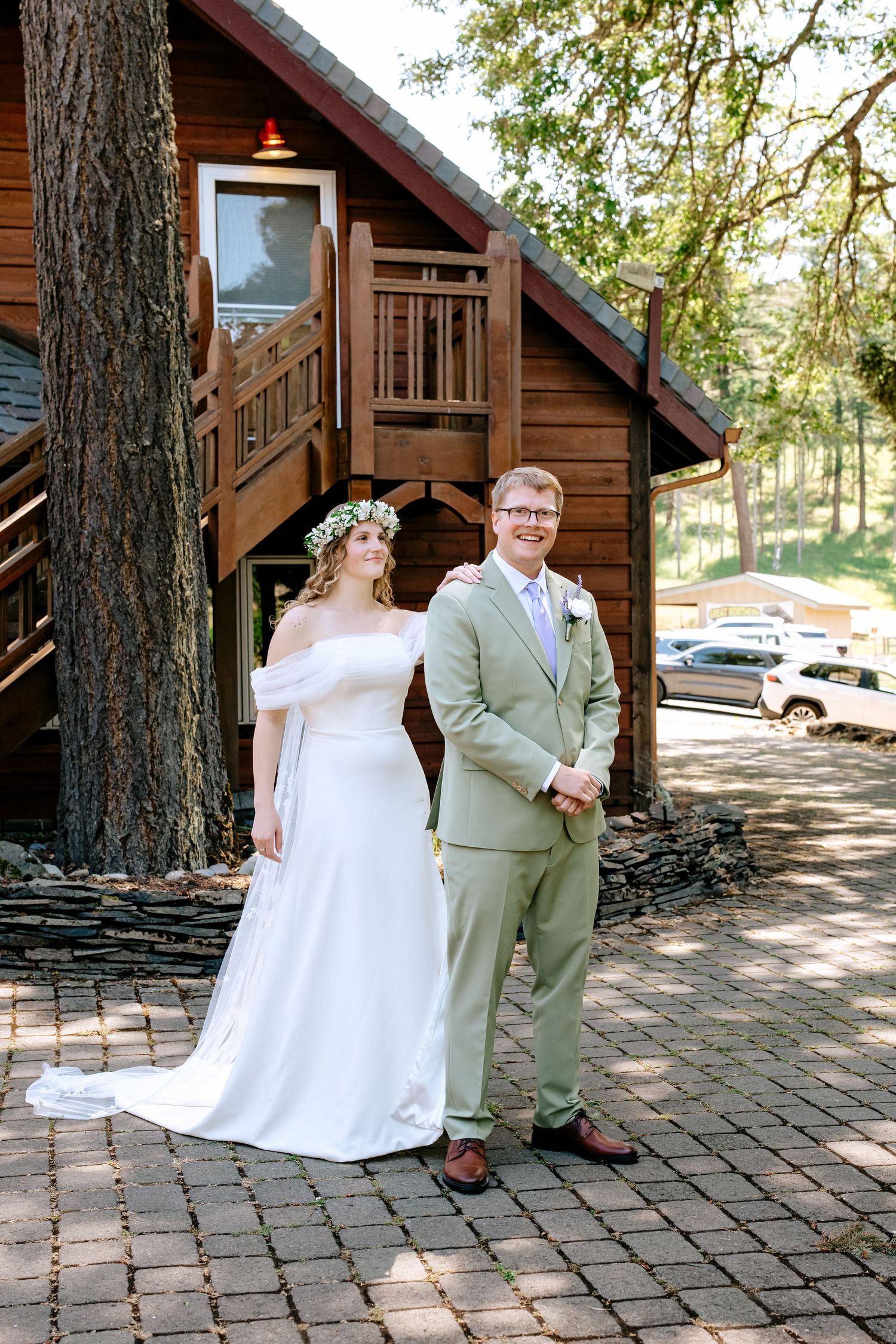 Bride in white gown with floral crown and groom in sage suit pose outside rustic wooden cabin on their wedding day.