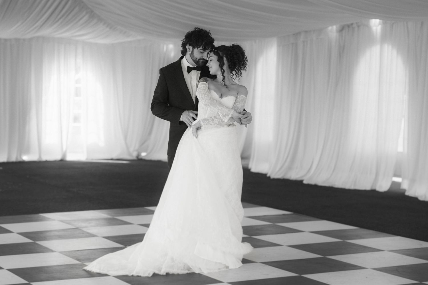 Black and white photo of a bride and groom sharing their first dance in an elegant white draped wedding venue.