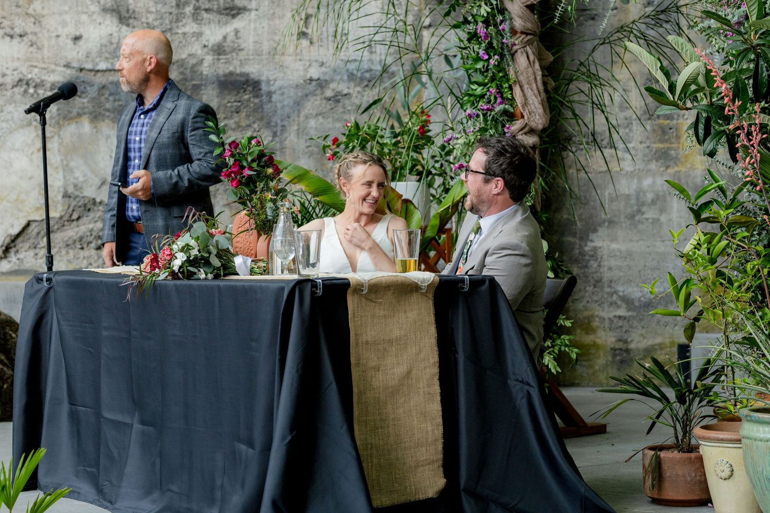 Bride and groom smile at sweetheart table with burlap runner while guest delivers speech at wedding reception.