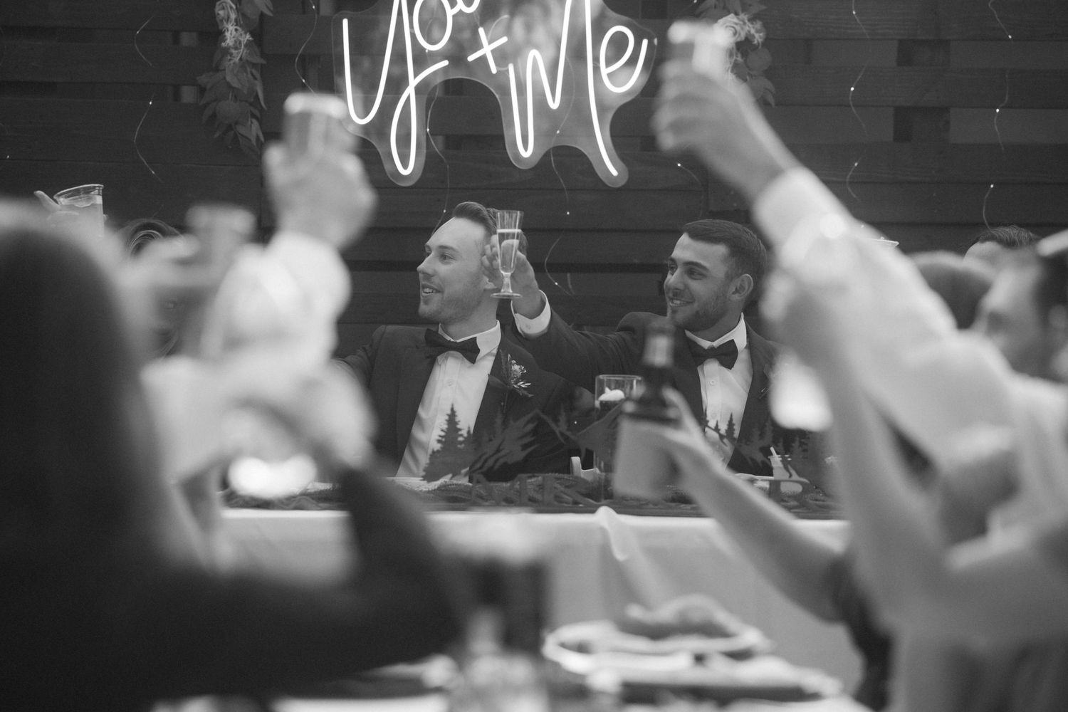 Black and white photo of two grooms raising glasses during a toast at their wedding reception.