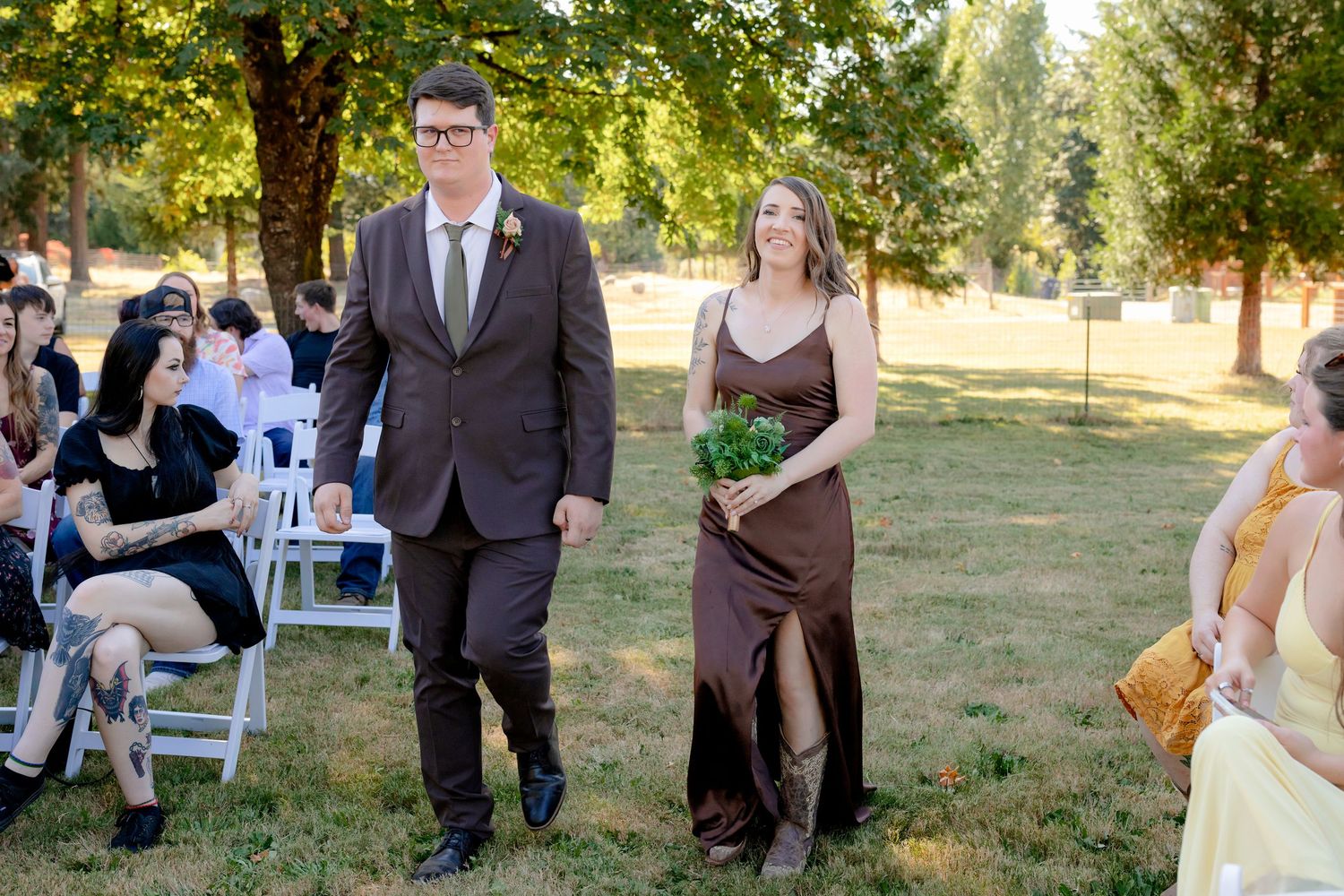 Groomsman in brown suit escorts bridesmaid in chocolate dress down outdoor wedding aisle.