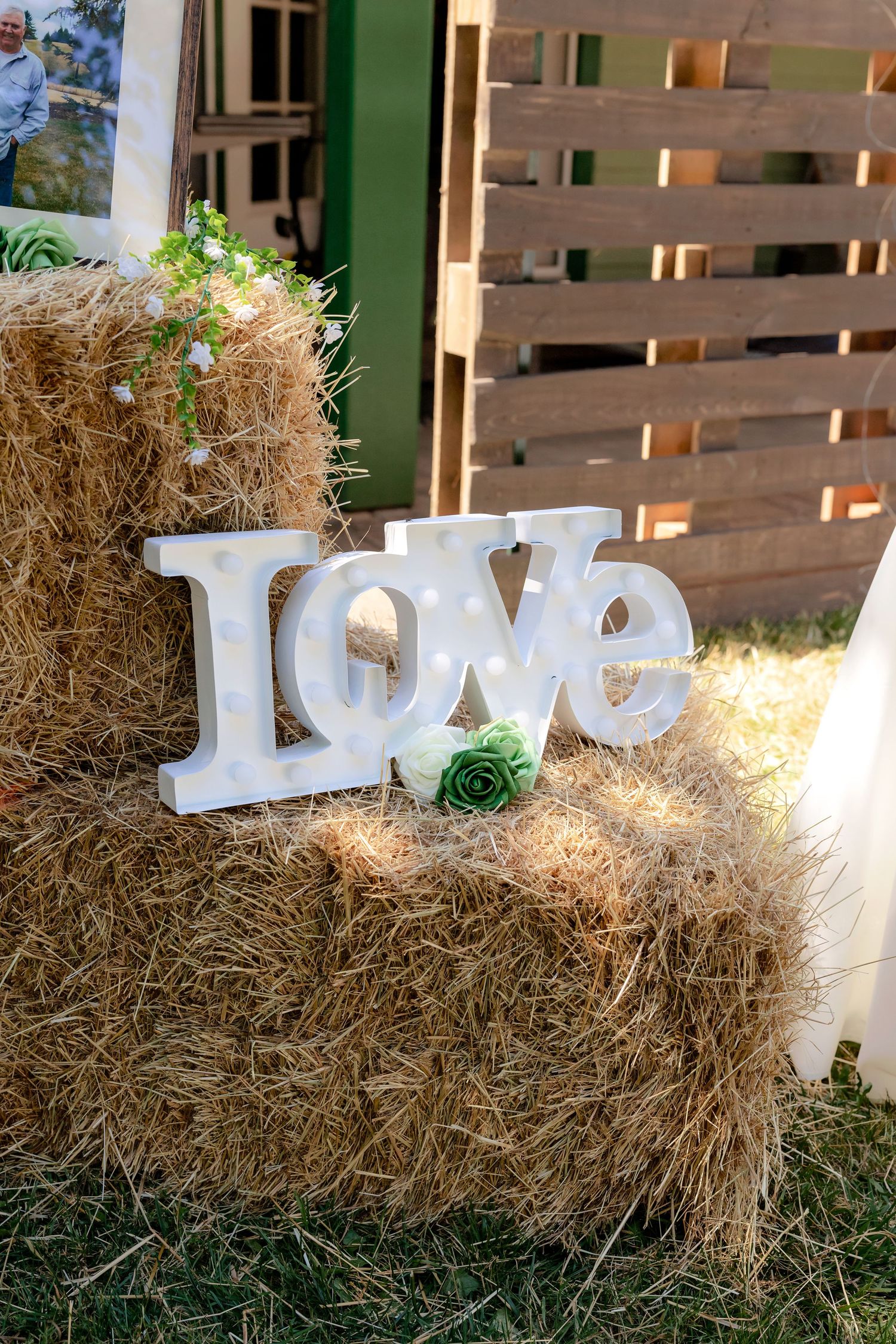 White marquee LOVE sign with green rose on rustic hay bale at outdoor wedding reception.