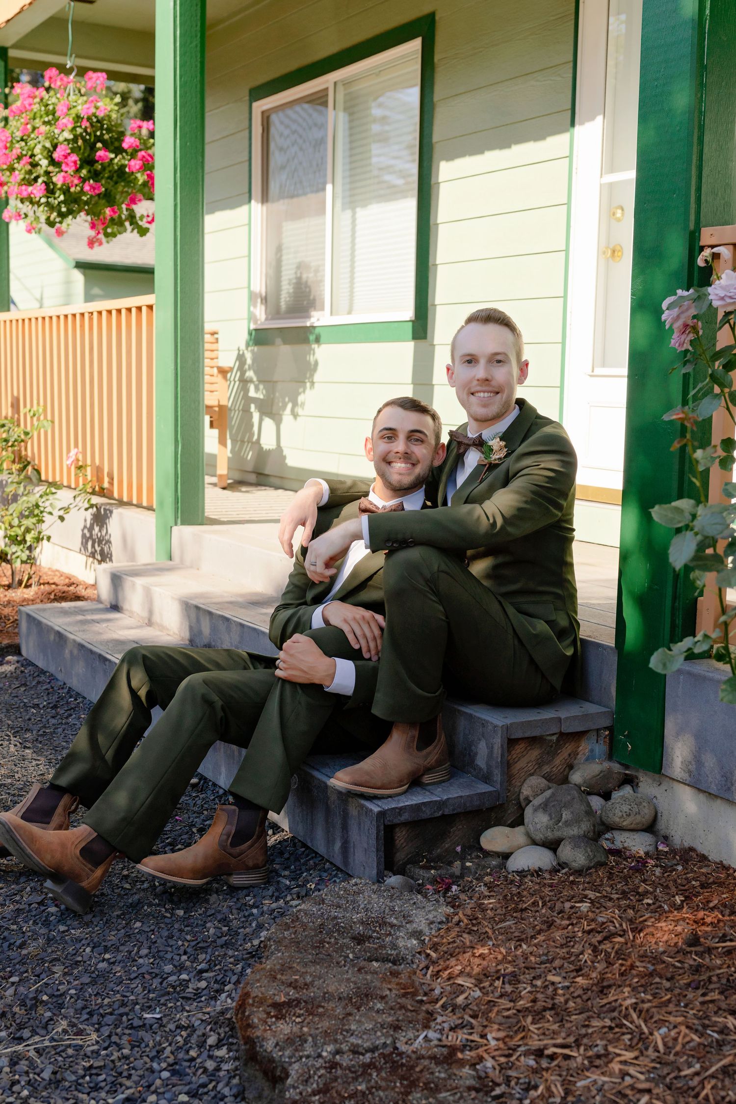 Two grooms in olive green suits sit together smiling on porch steps of a green house.