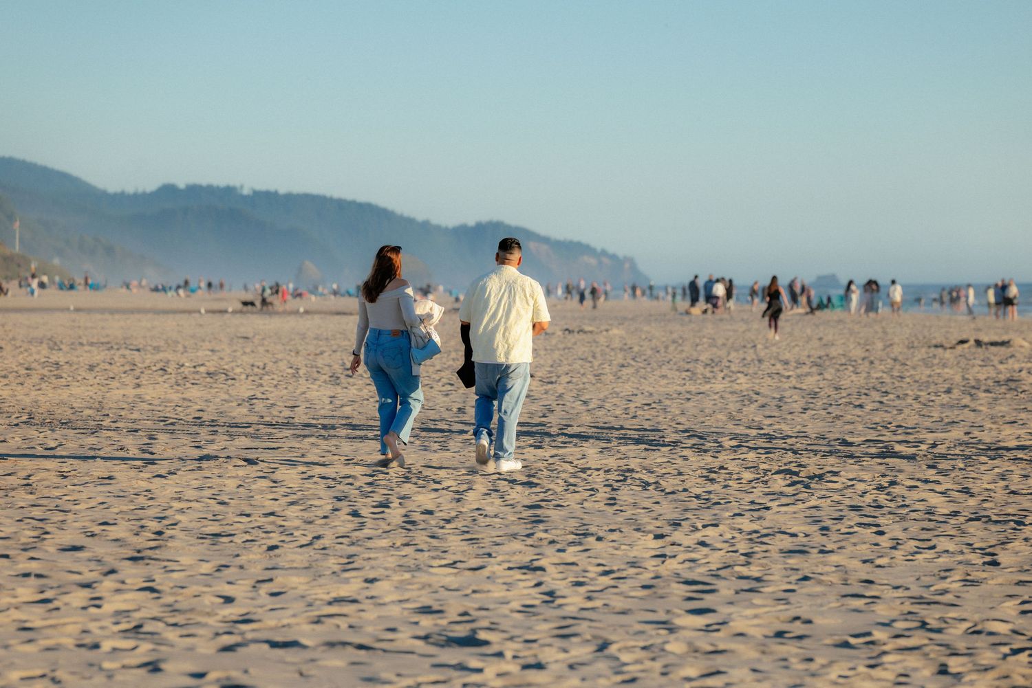 Two people walking hand in hand on a sandy beach with mountains and ocean in the background on a sunny day.