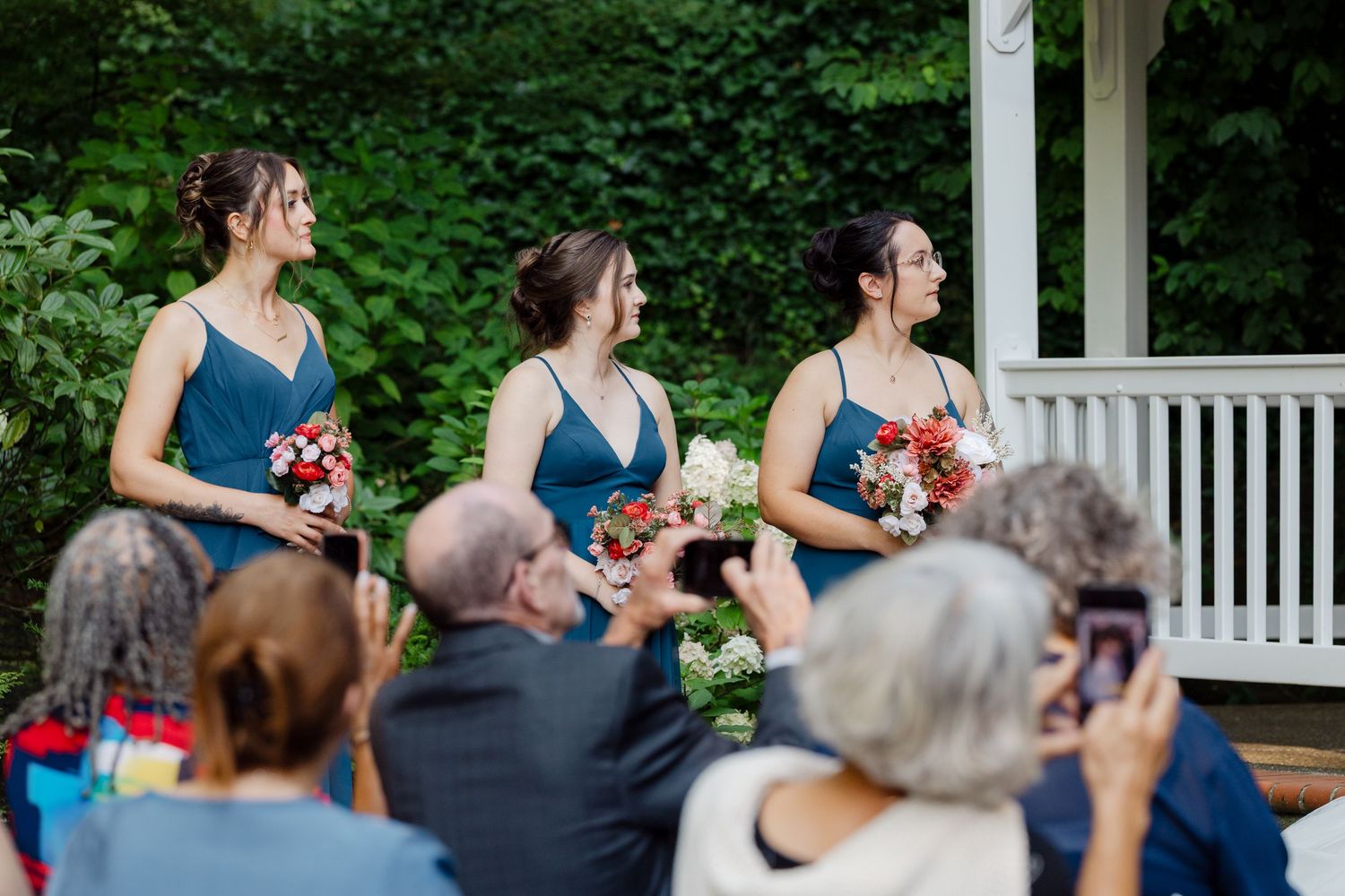 Three bridesmaids in blue dresses holding bouquets stand at outdoor wedding ceremony before seated guests.