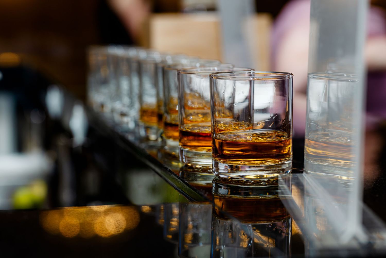 Row of whiskey glasses filled with amber spirits lined up on a reflective bar surface.
