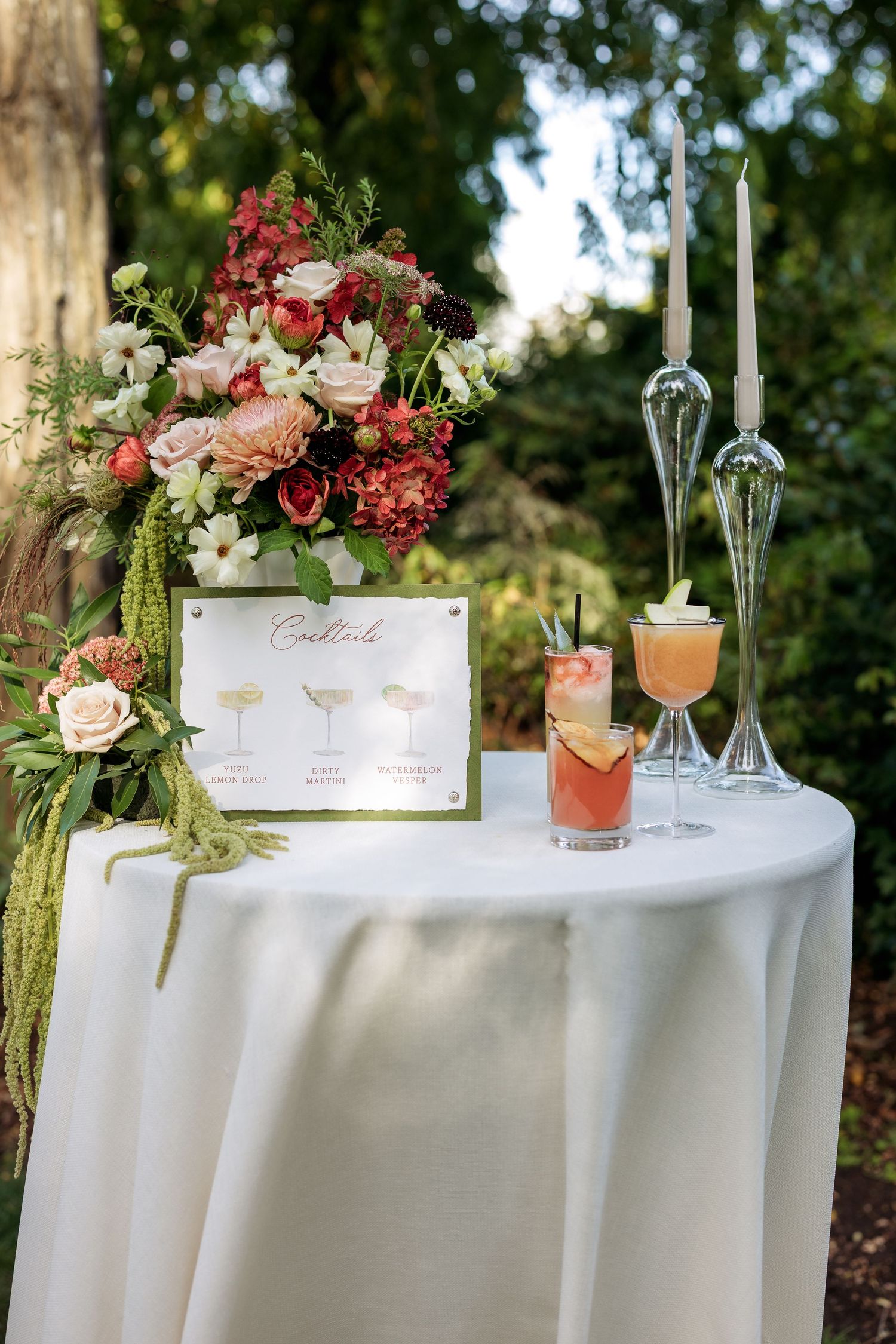 Elegant wedding cocktail table with white linens, floral arrangements, and crystal glassware set in an outdoor garden setting.