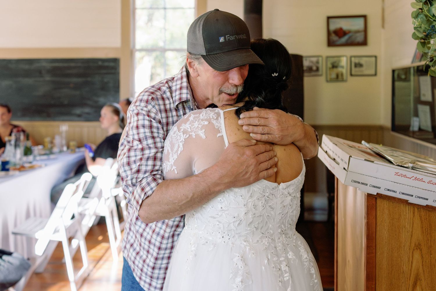 An emotional embrace between two people at an indoor venue, with one wearing a plaid shirt and baseball cap.