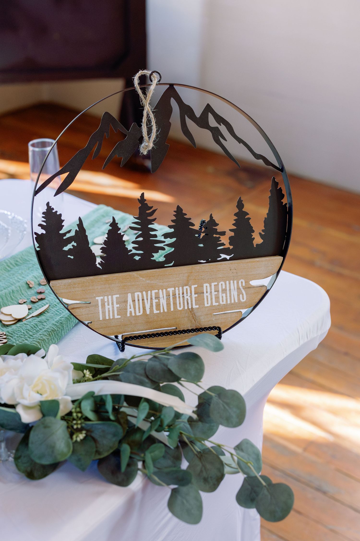 A rustic wedding sign reading 'The Adventure Begins' decorated with pine tree silhouettes and greenery.