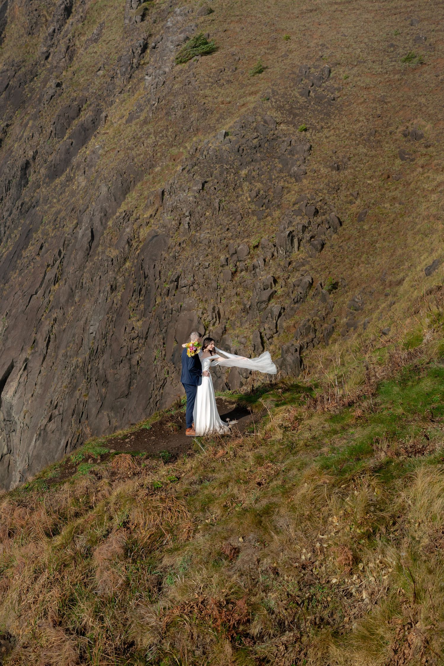 A couple in wedding attire poses dramatically on a steep mountainside cliff against a dramatic natural backdrop.