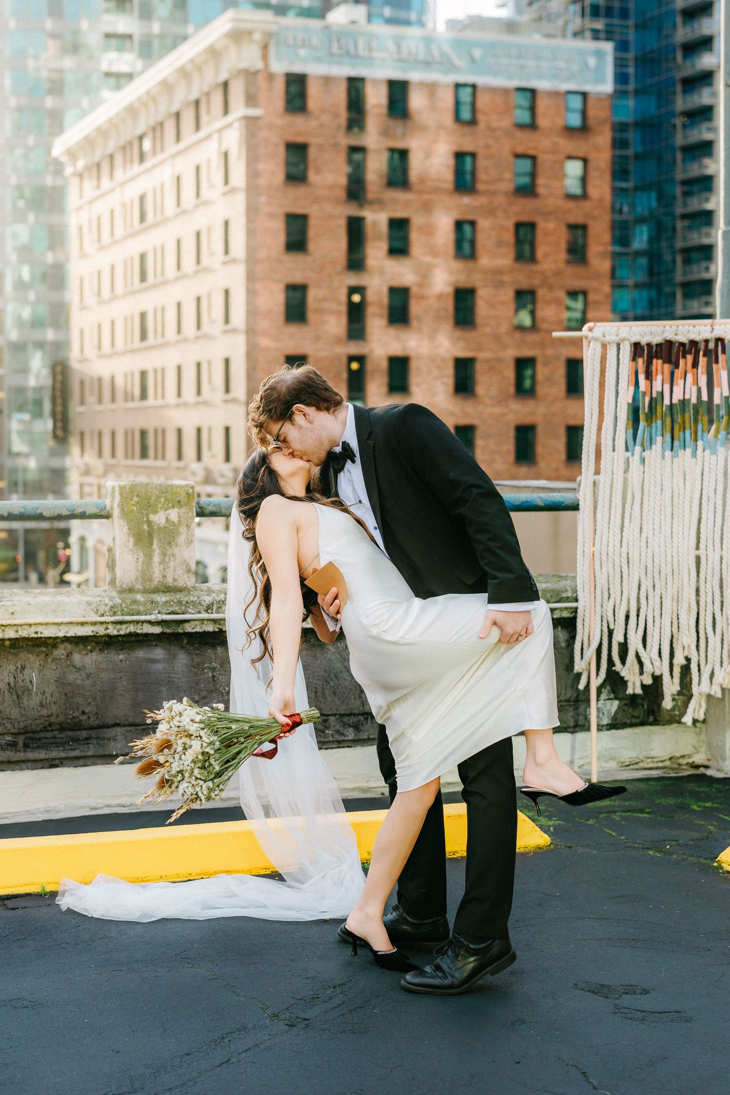 Couple performing romantic dip pose on urban rooftop with brick buildings behind.