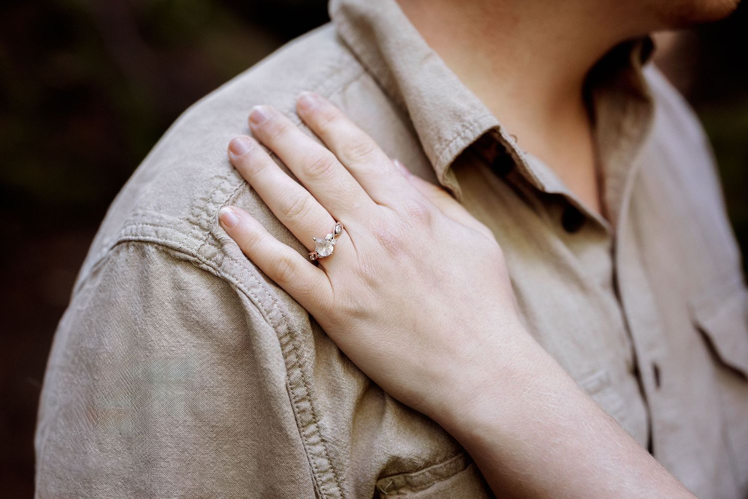 A close-up shot of a hand with an engagement ring resting on a beige shirt shoulder.