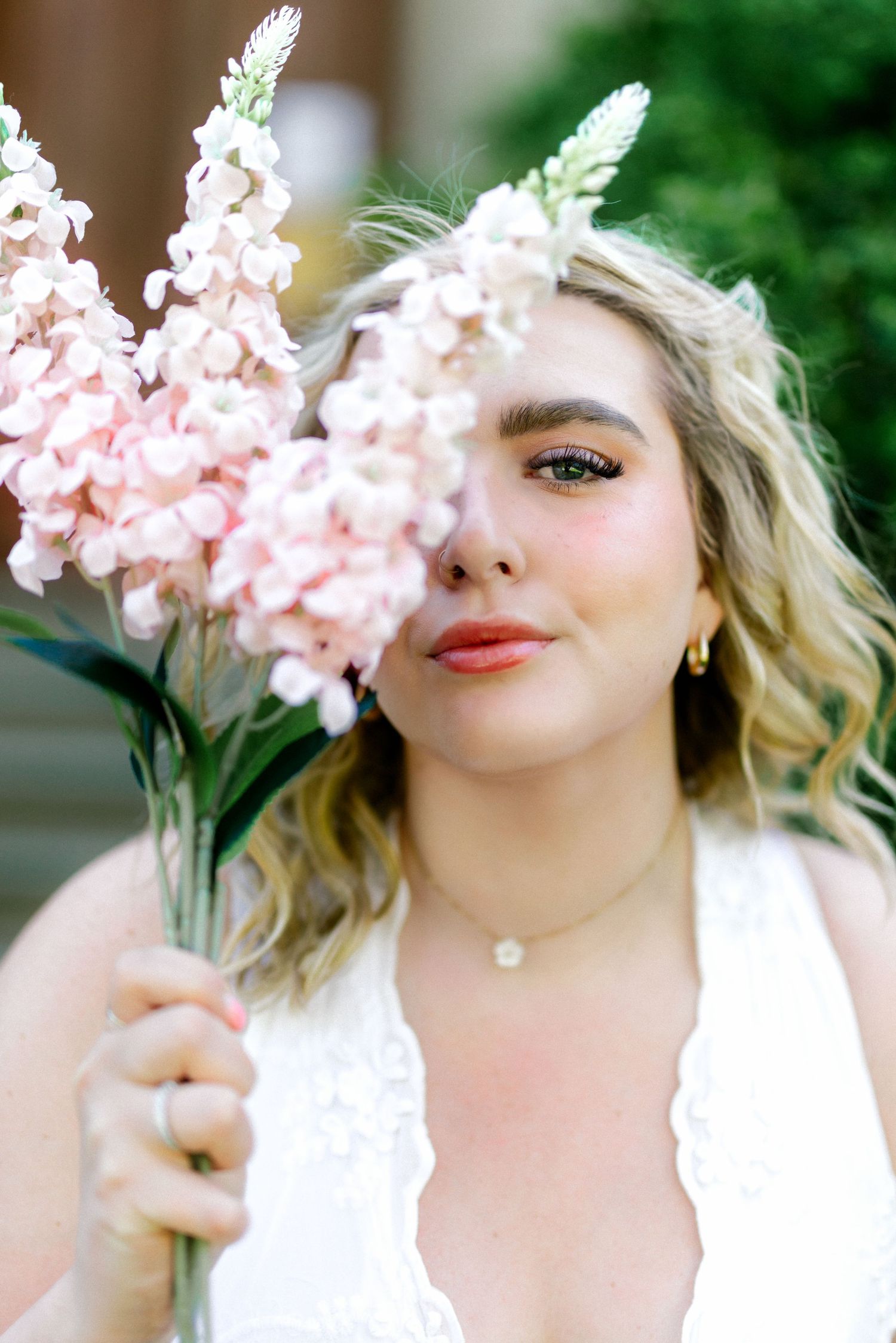 Blonde woman in white dress holding pink snapdragon flowers partially covering her face, smiling softly outdoors.