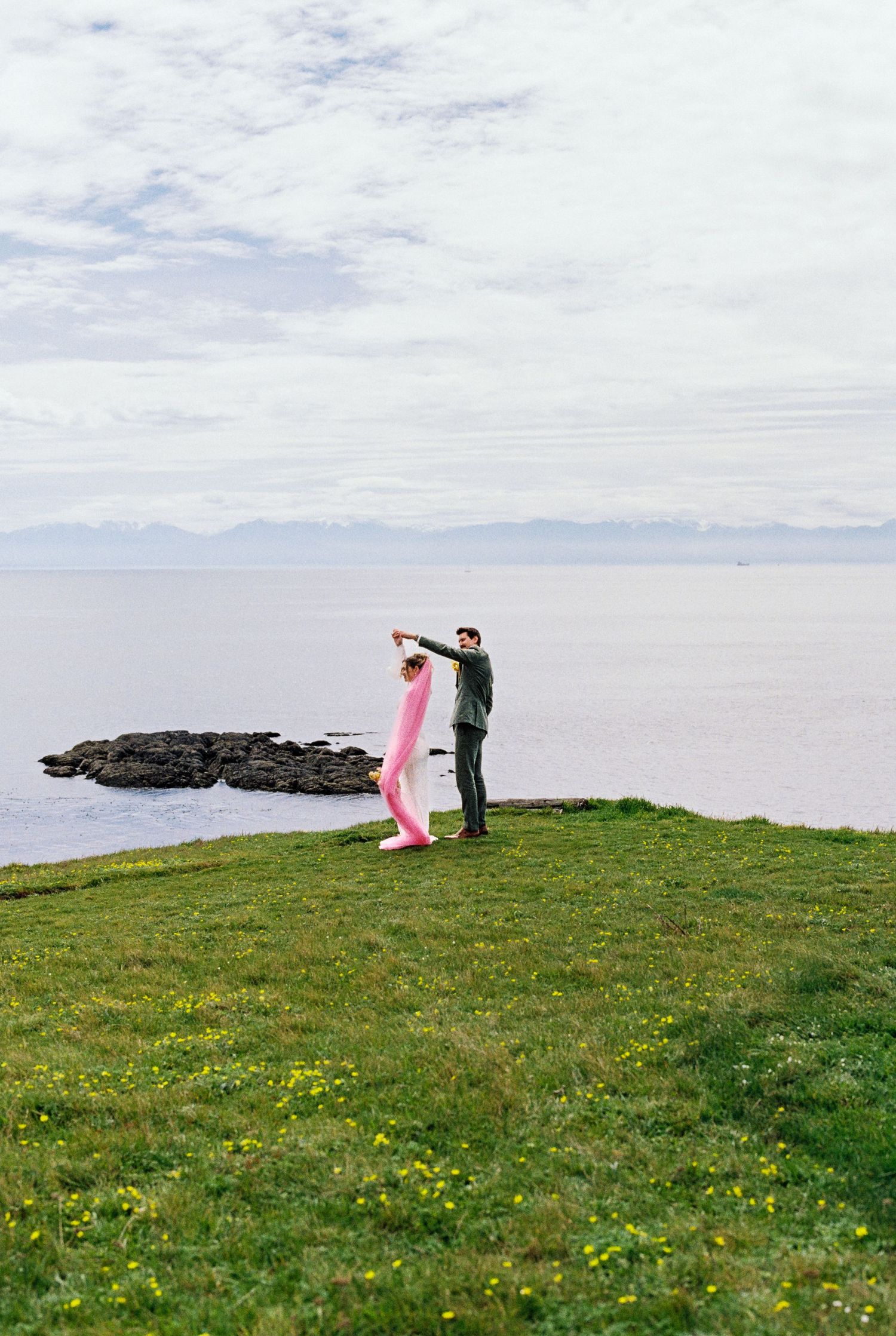 Bride in pink outfit and groom in dark suit dance joyfully on grassy coastal cliff overlooking calm ocean.