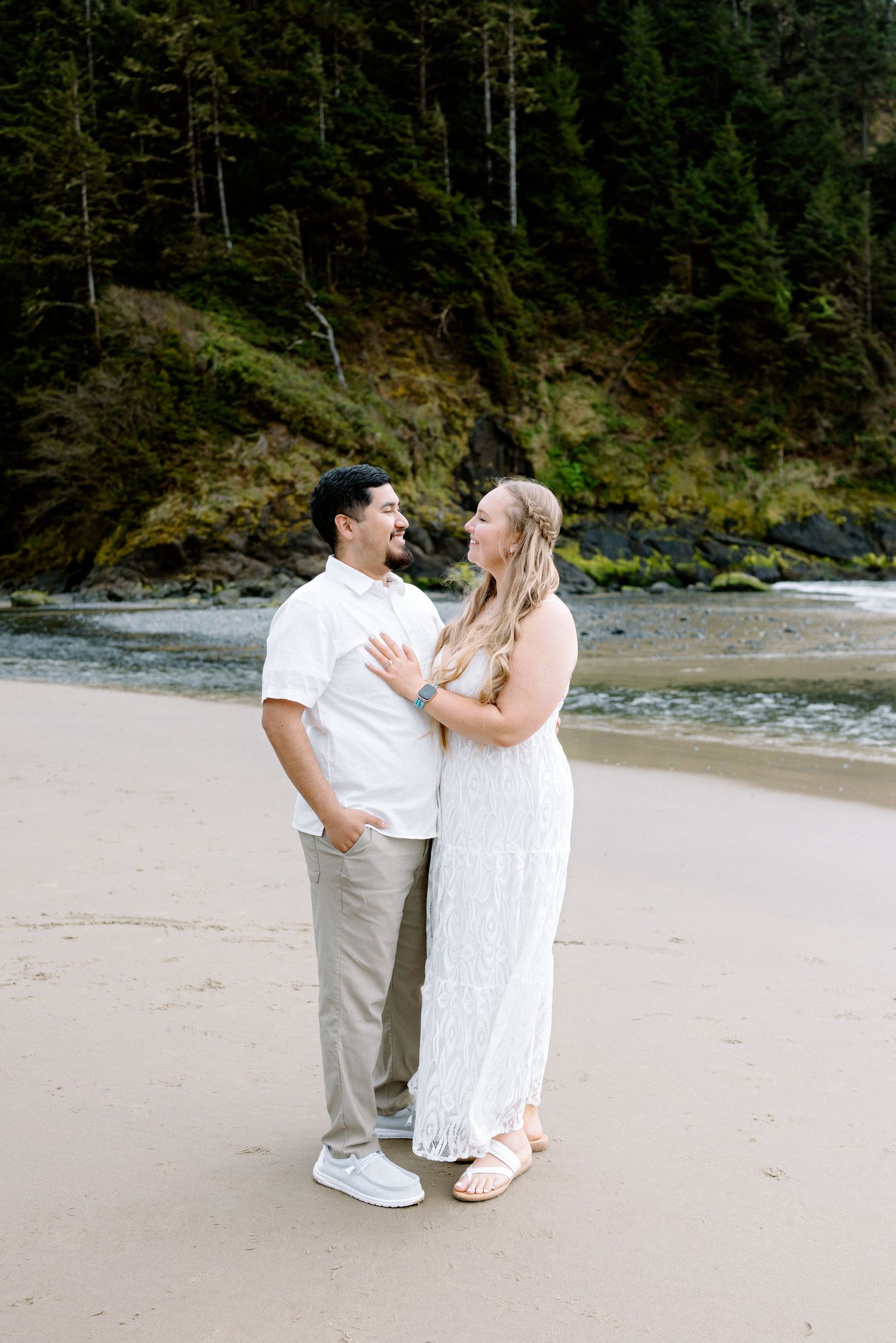Couple in white outfits embracing on sandy beach with lush green forest and rocky cliffs in background.