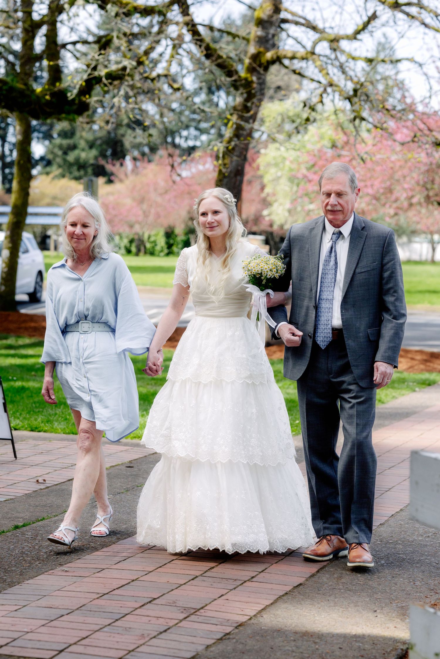 Bride in white lace gown walks down aisle escorted by parents at outdoor spring wedding ceremony.