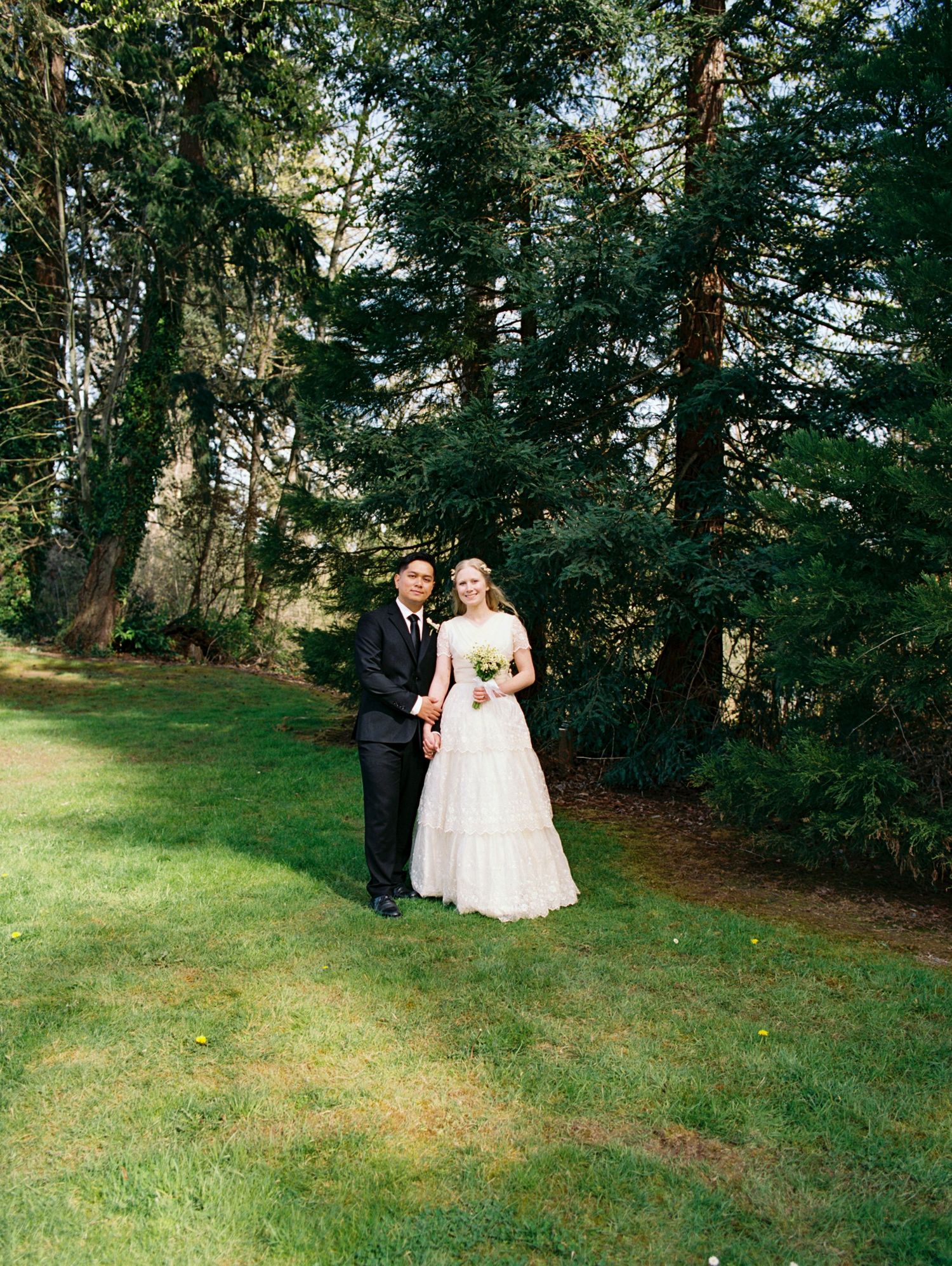 Bride and groom pose together on green lawn surrounded by tall evergreen trees after wedding.