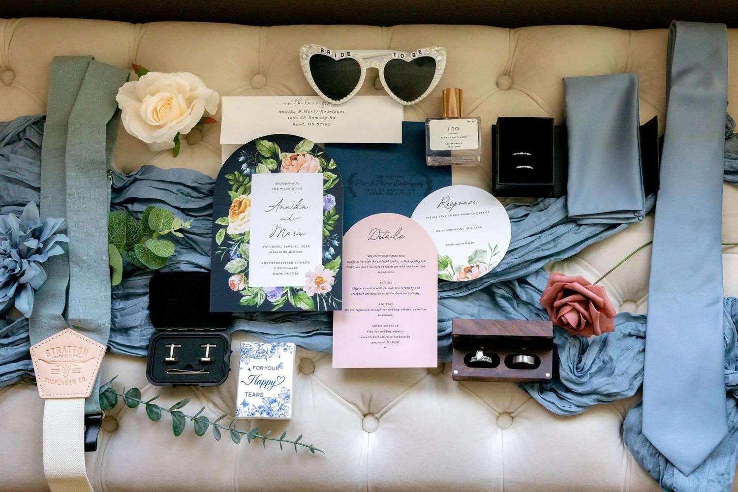 Elegant wedding flat lay featuring floral invitations, dusty blue ties, ring boxes, and heart sunglasses on a cream surface.
