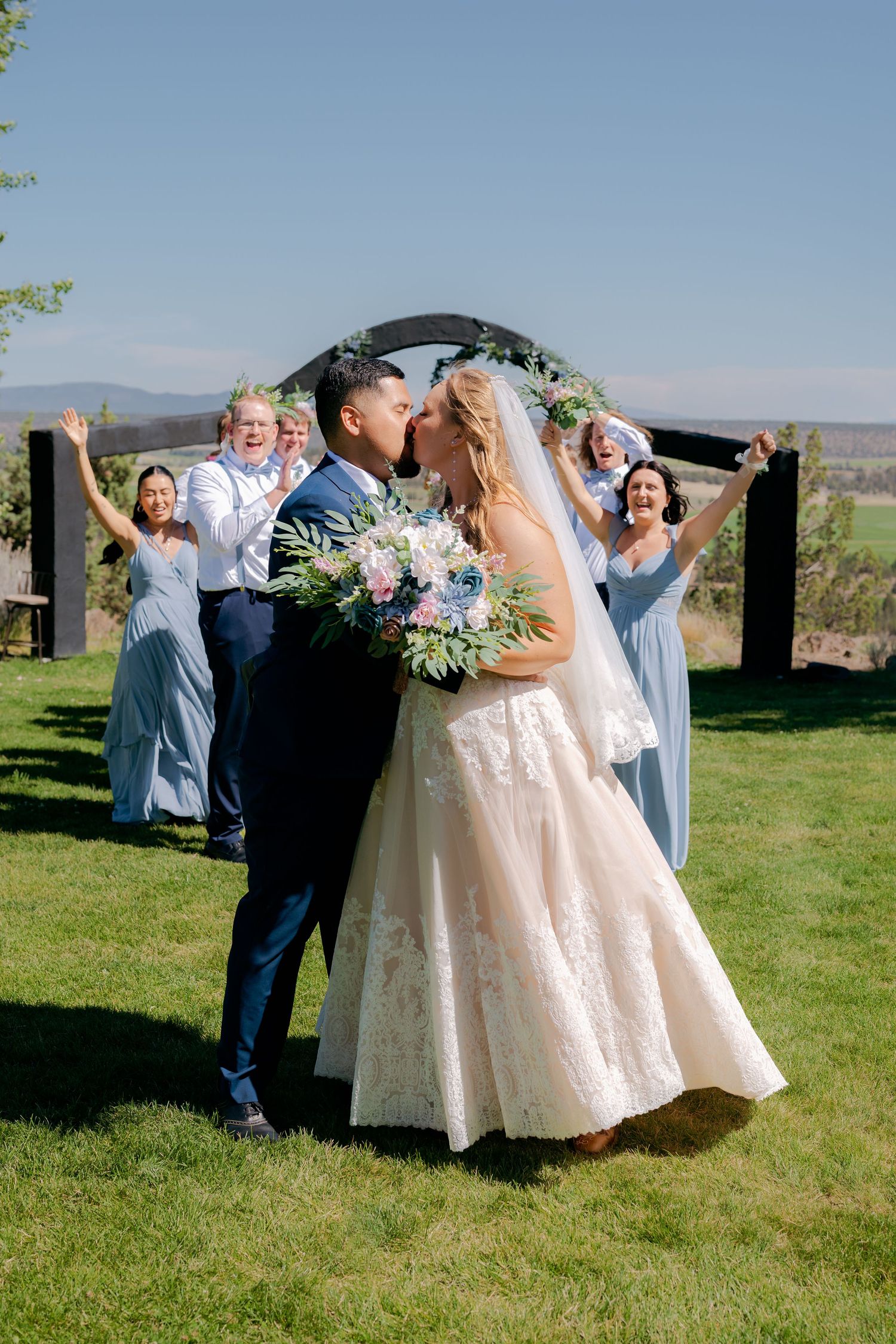 Newlyweds share first kiss under circular floral arch as wedding party cheers behind them outdoors.
