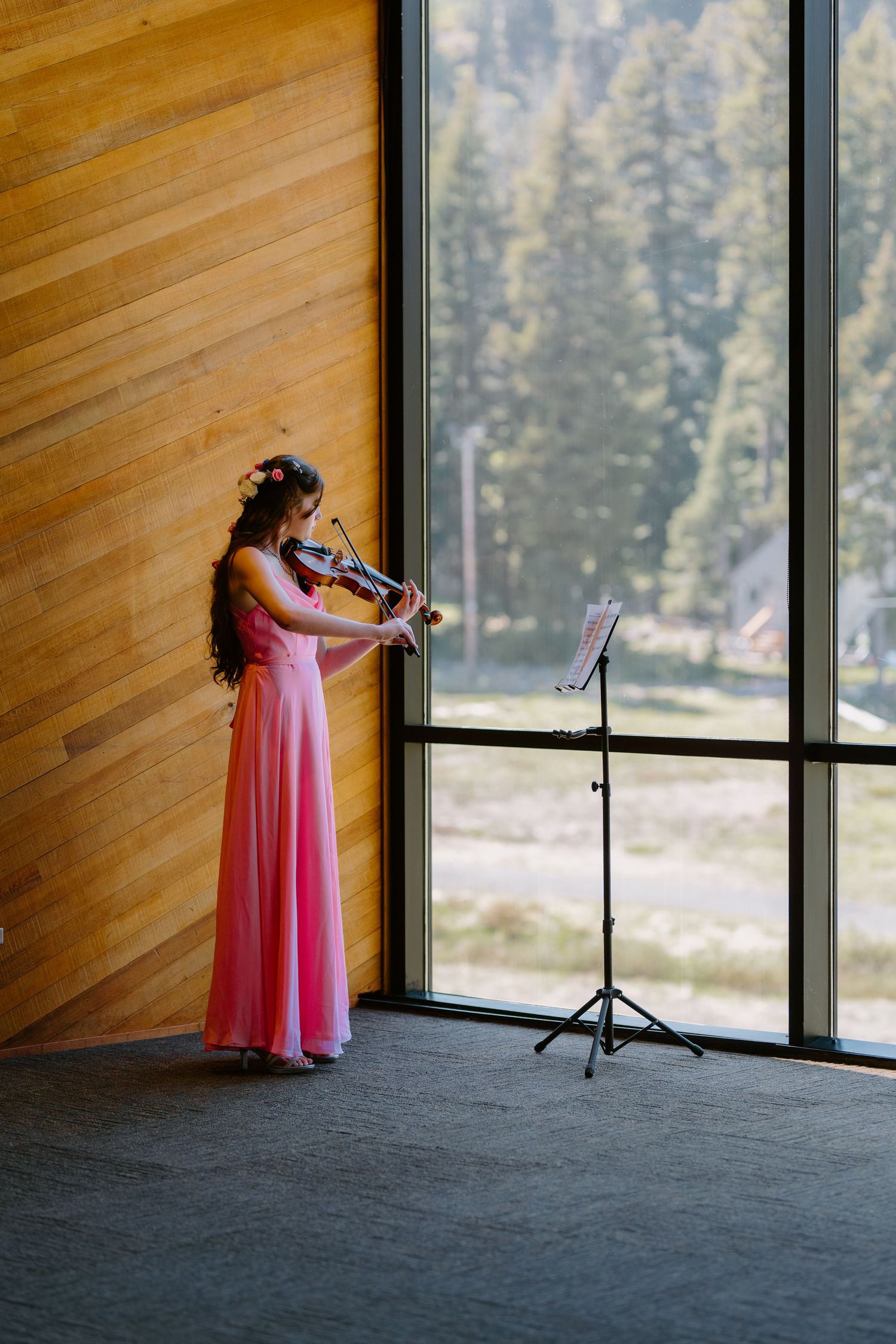 Violinist in pink gown performing beside large windows with forest view at wedding ceremony.