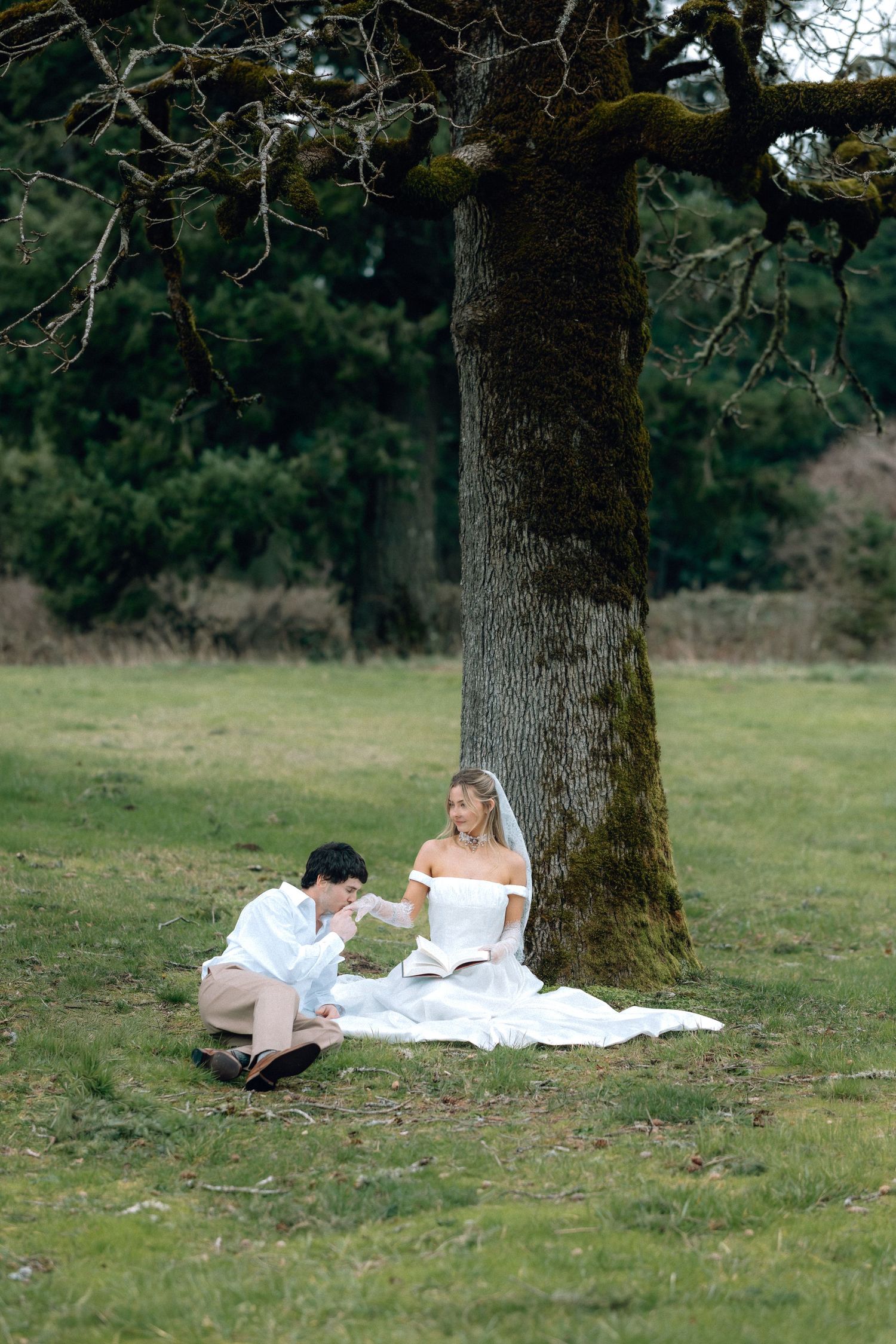 Couple relaxing on a white blanket under a large tree in a peaceful meadow setting.