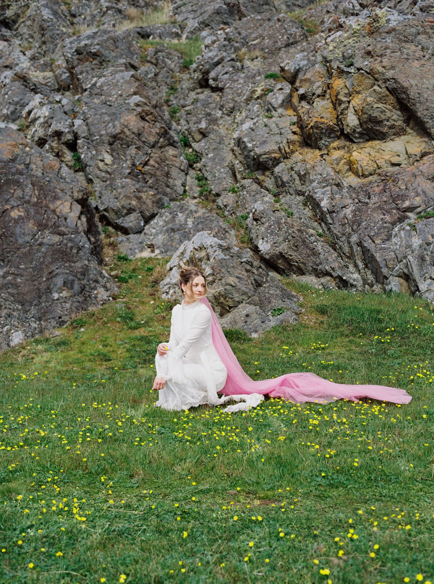 Bride in white wedding gown with flowing pink veil sits gracefully in yellow wildflower meadow before rocky cliffs.