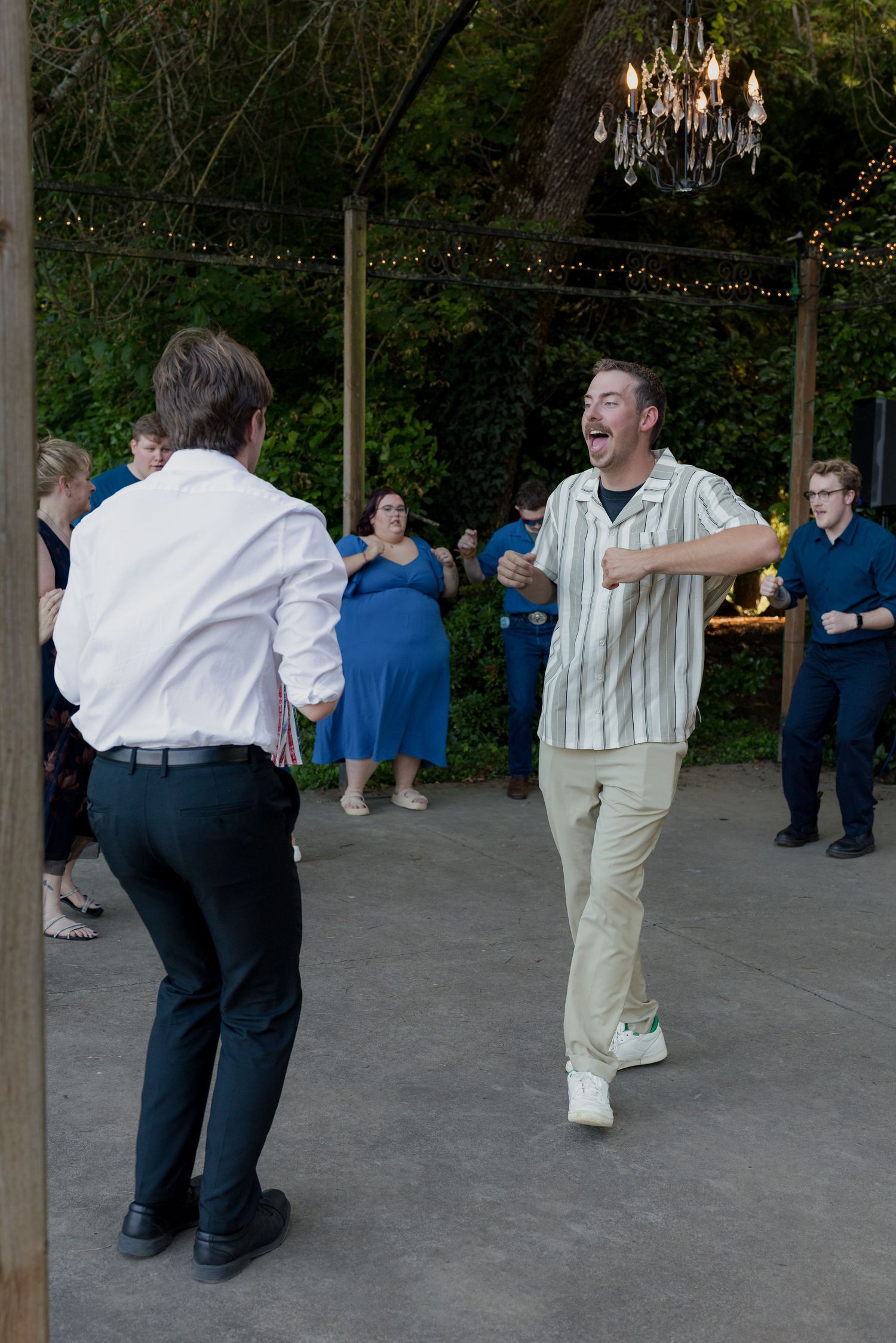 Two men dance energetically at an outdoor wedding reception under string lights at night.
