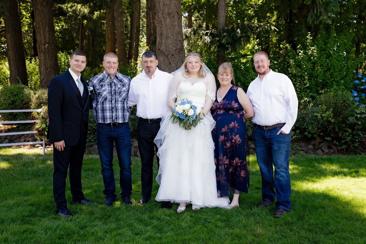 Wedding party poses outdoors on grass with tall evergreen trees in background, bride in white gown holding blue bouquet.