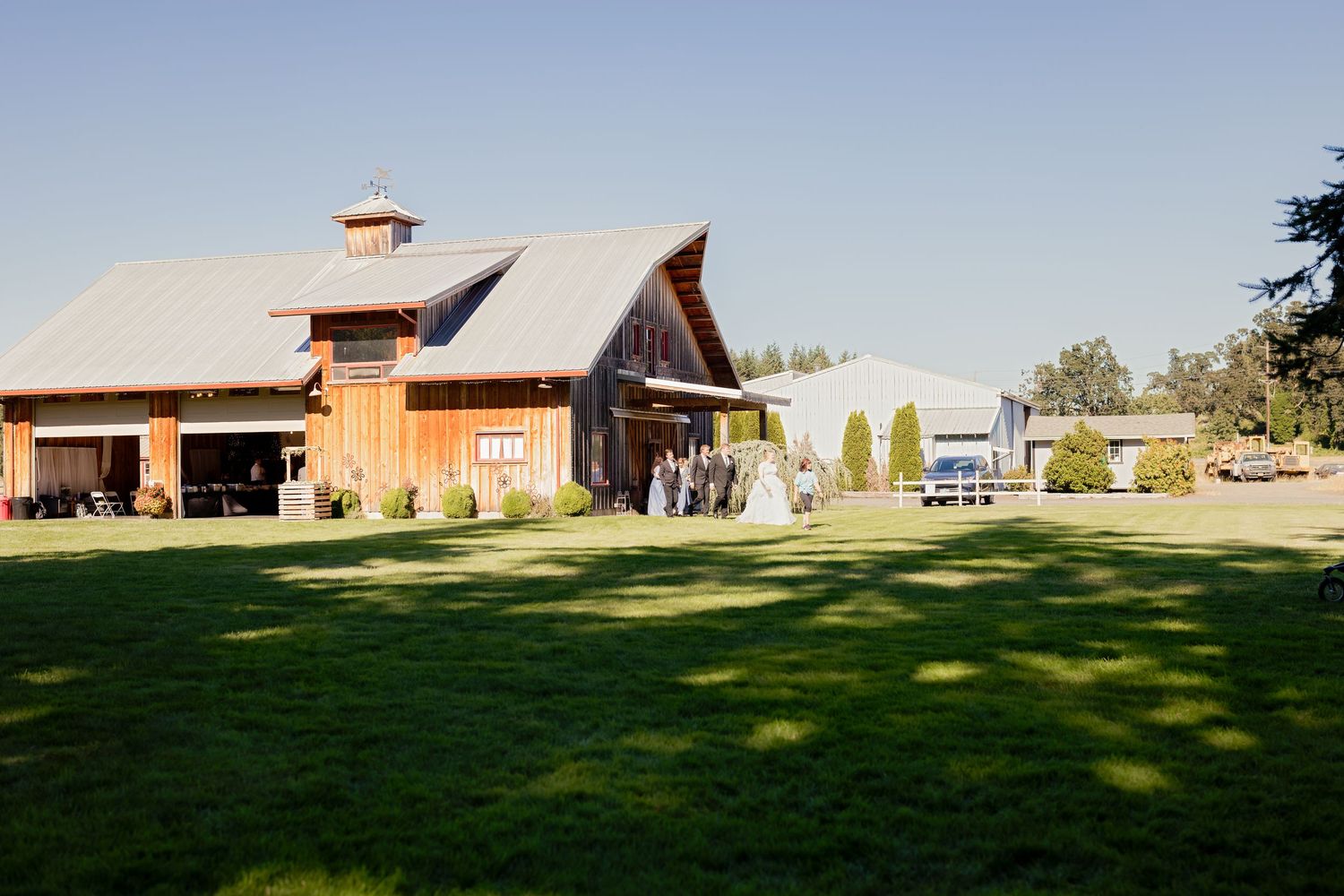 Rustic barn wedding venue with large green lawn and wedding party gathered near entrance on sunny day.