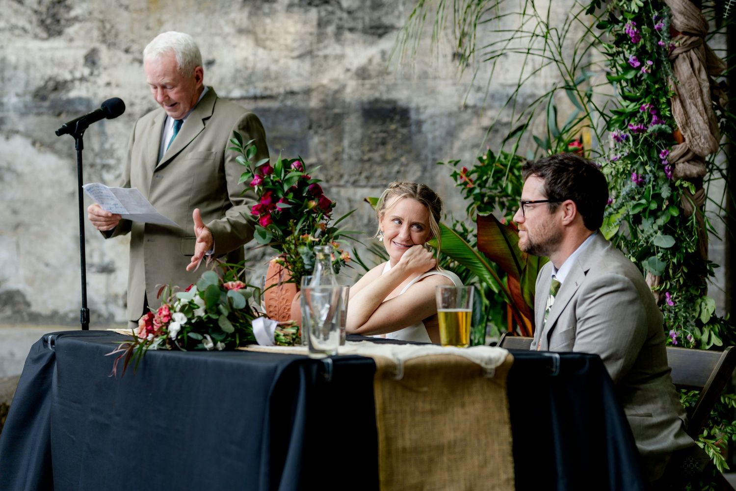 Elderly man giving a wedding speech at a reception while the bride and groom smile at a decorated table.